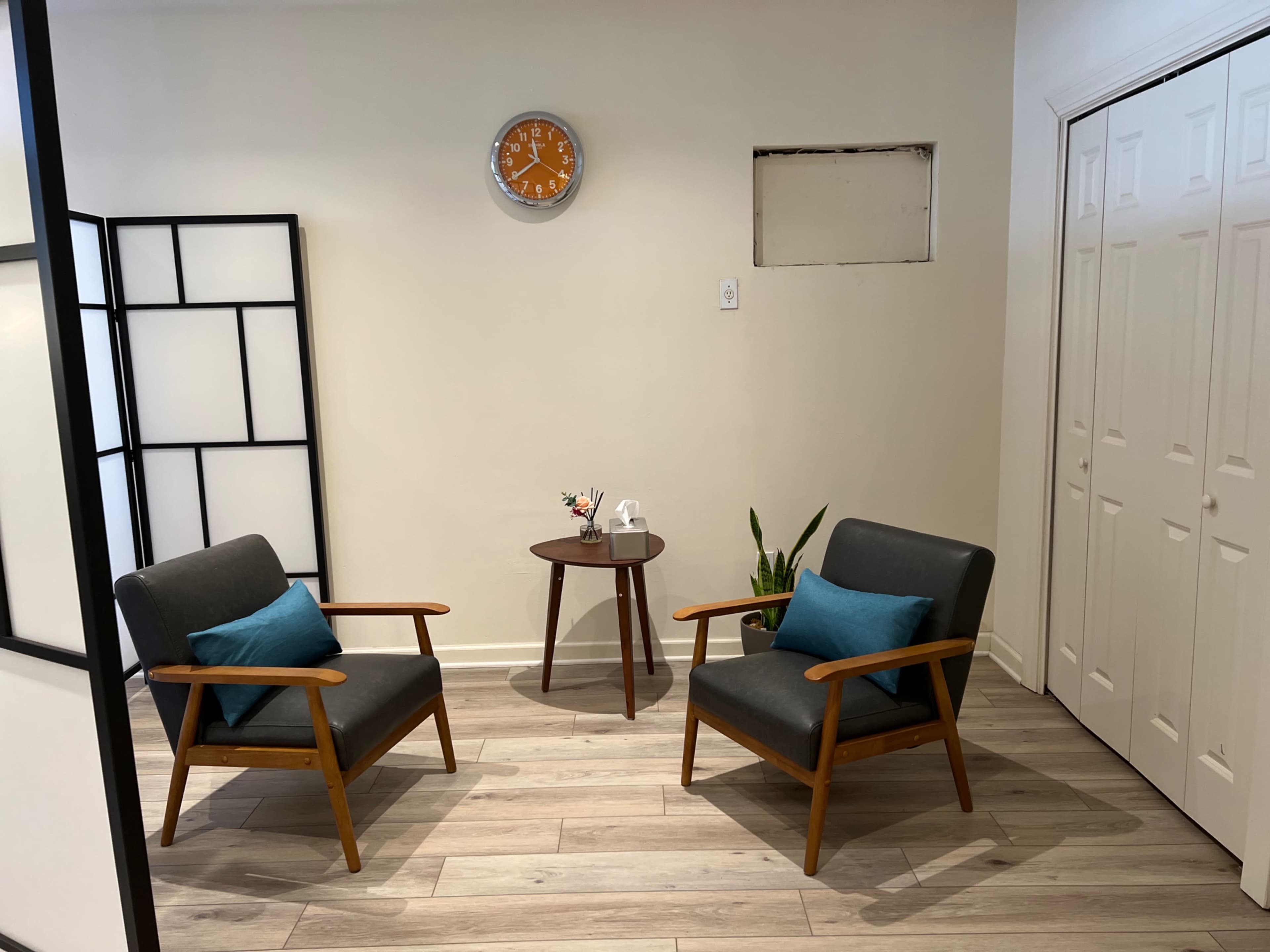A spacious room features two gray chairs with wooden arms, a small round table between them, and a wall clock above, surrounded by light-colored flooring.