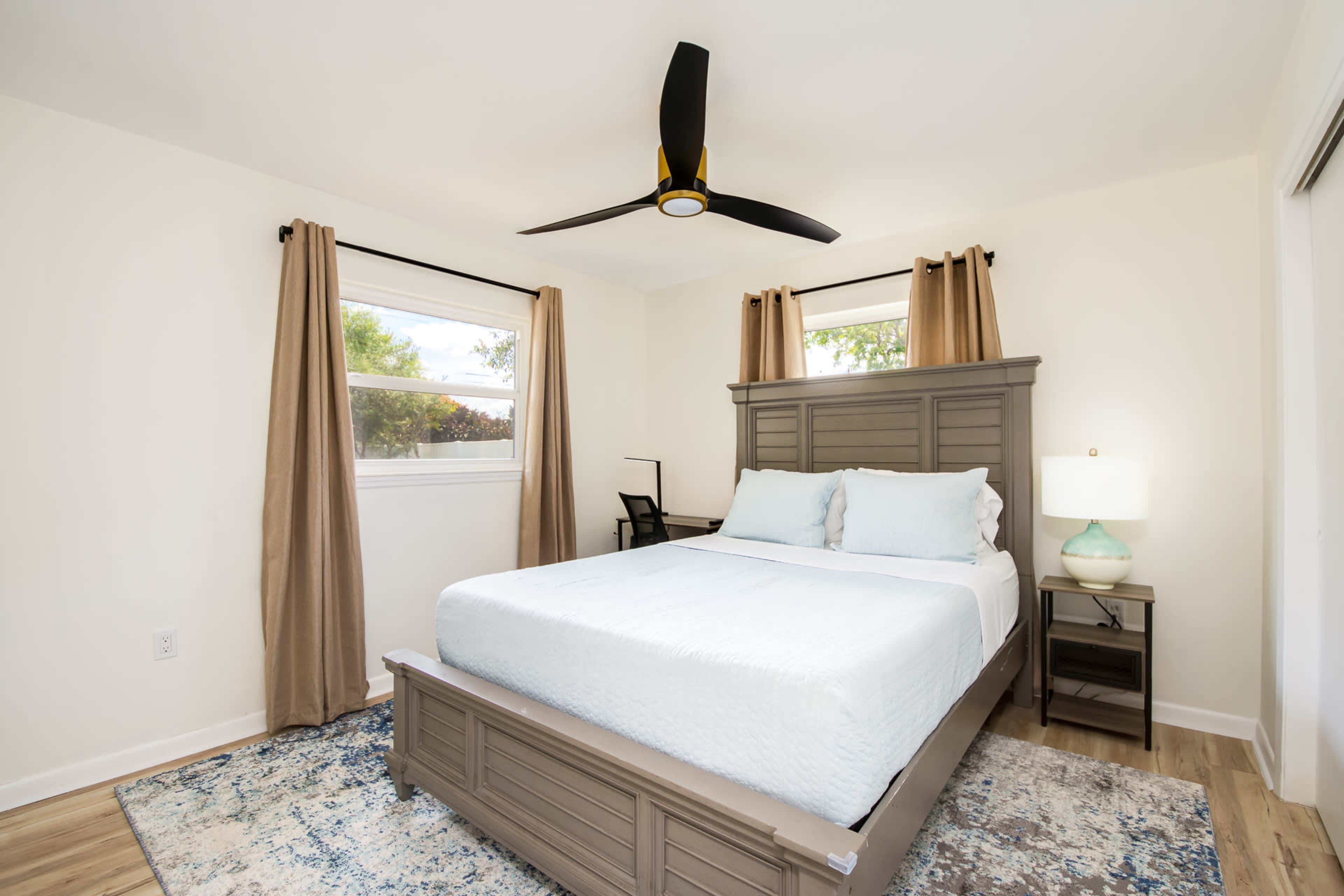 A neatly arranged bedroom with a large bed, two windows, a ceiling fan, and a bedside table with a lamp.