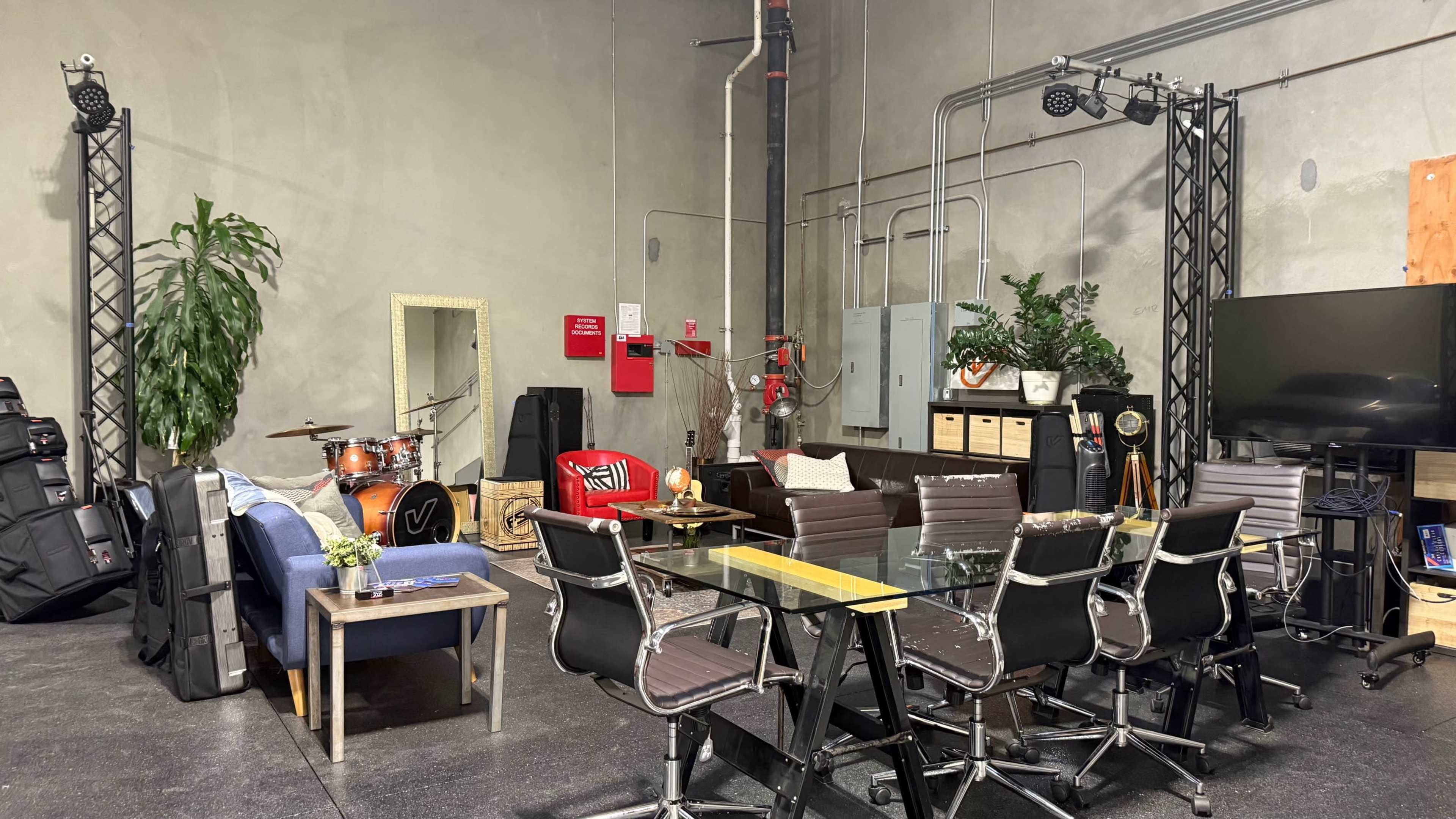 The image shows a spacious room with a seating area featuring a couch and chairs, a dining table, musical instruments, plants, and various storage units.