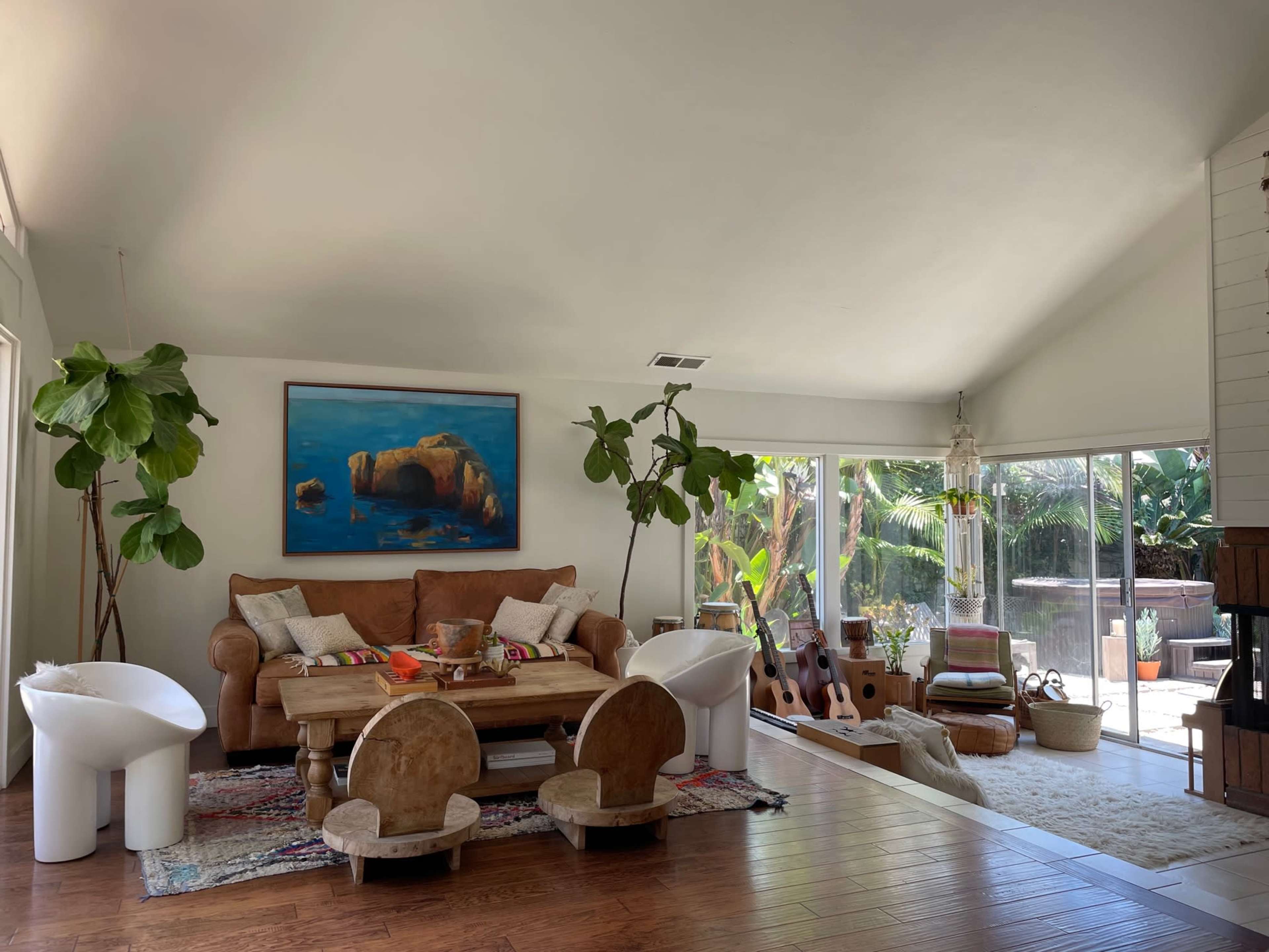The image depicts a living room with a large brown sofa, a wooden table, and various plants, surrounded by large windows that offer a view of the outdoors.
