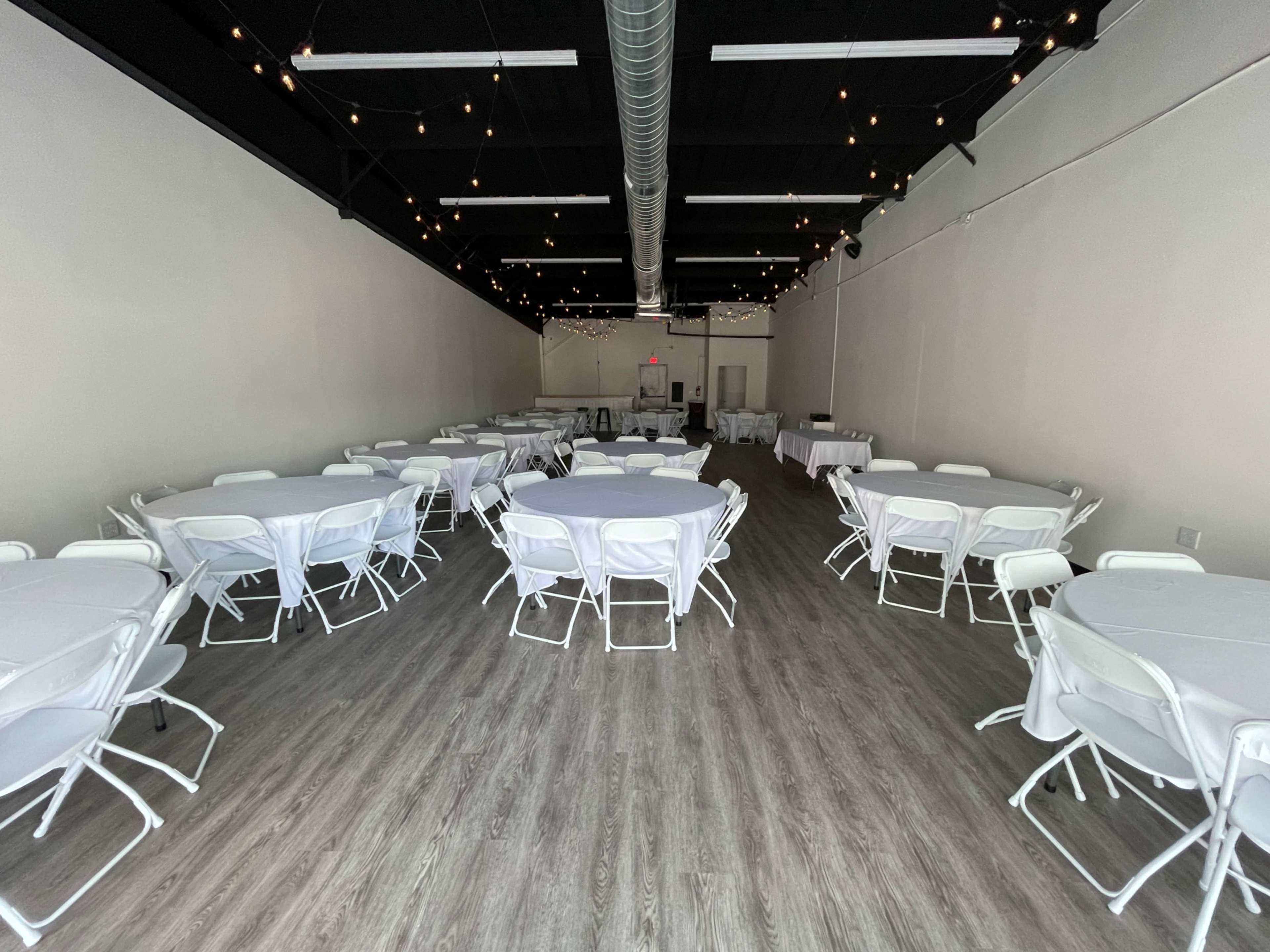 A spacious event room features several round tables covered with white tablecloths, surrounded by white folding chairs, and is illuminated by string lights overhead.