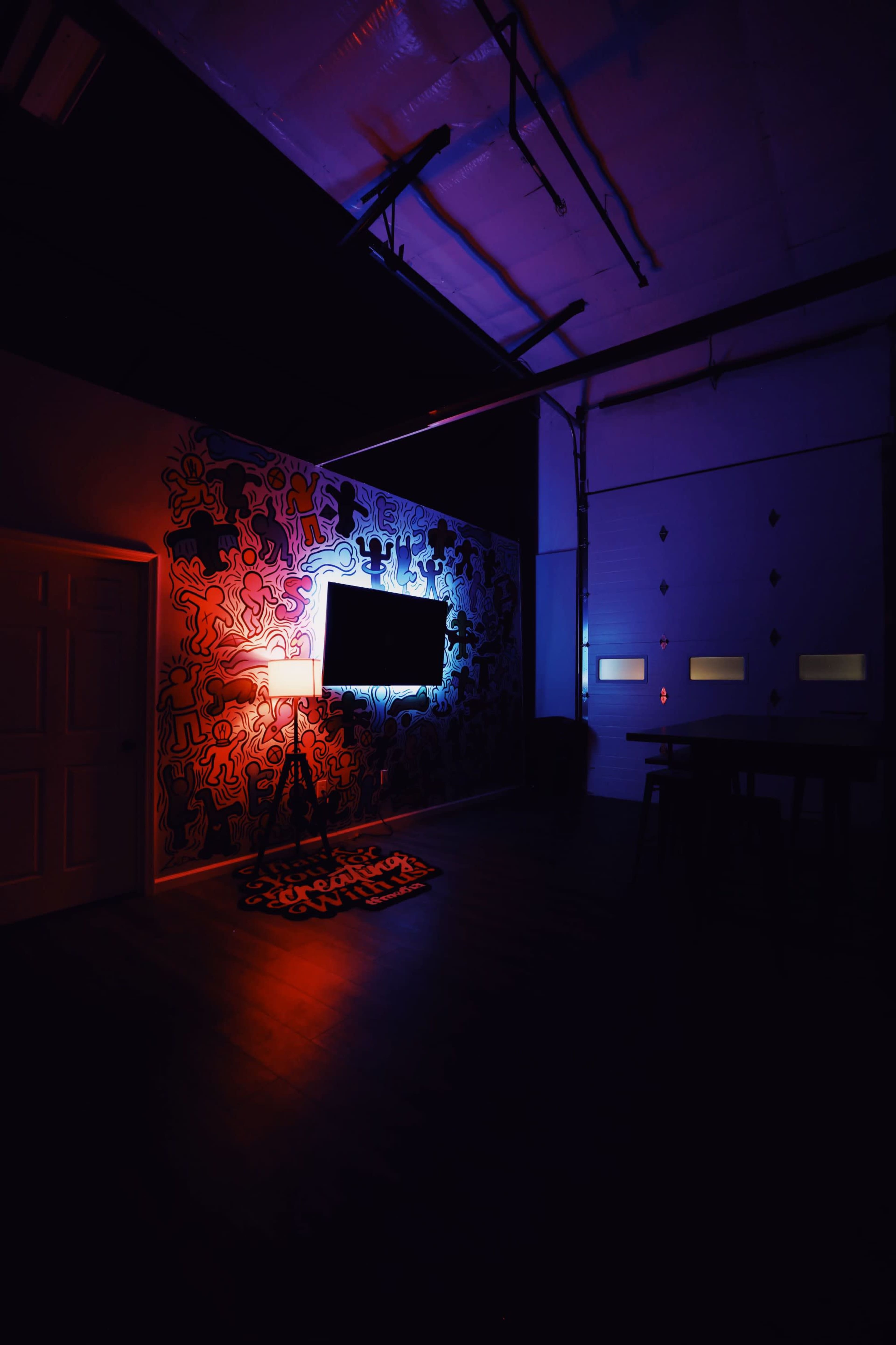 A dimly lit room with a colorful wall mural, a television mounted on the wall, and a floor lamp casting light on a patterned rug.