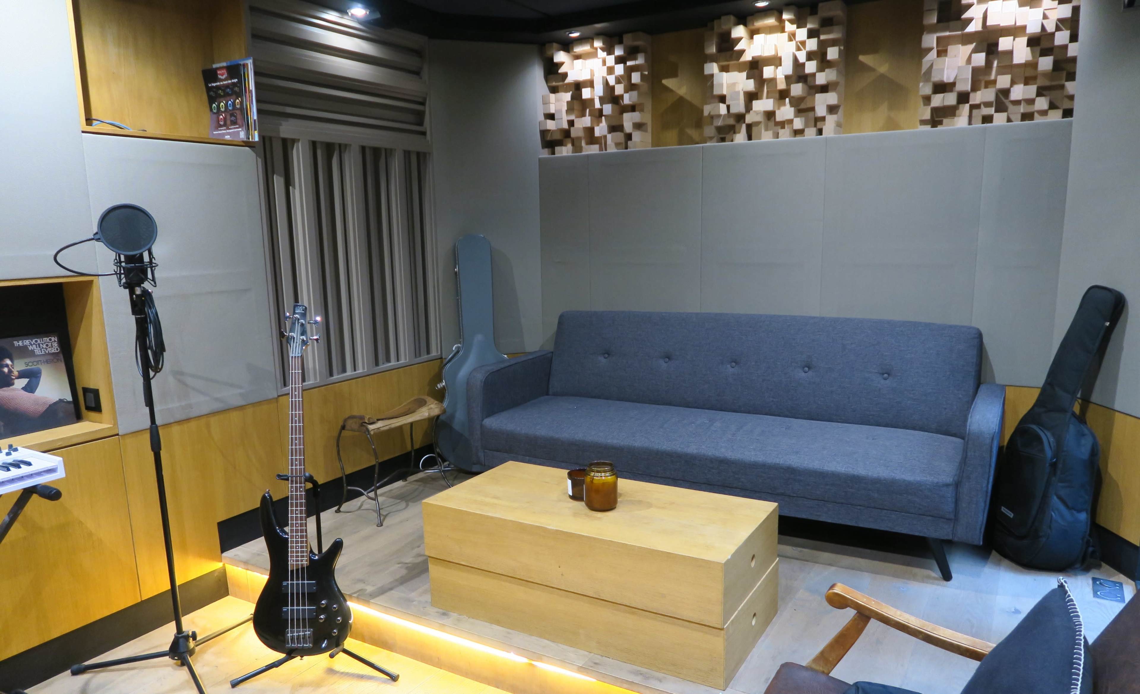 The image shows a music recording room featuring a gray sofa, a microphone, a bass guitar, and wooden soundproofing panels on the walls.