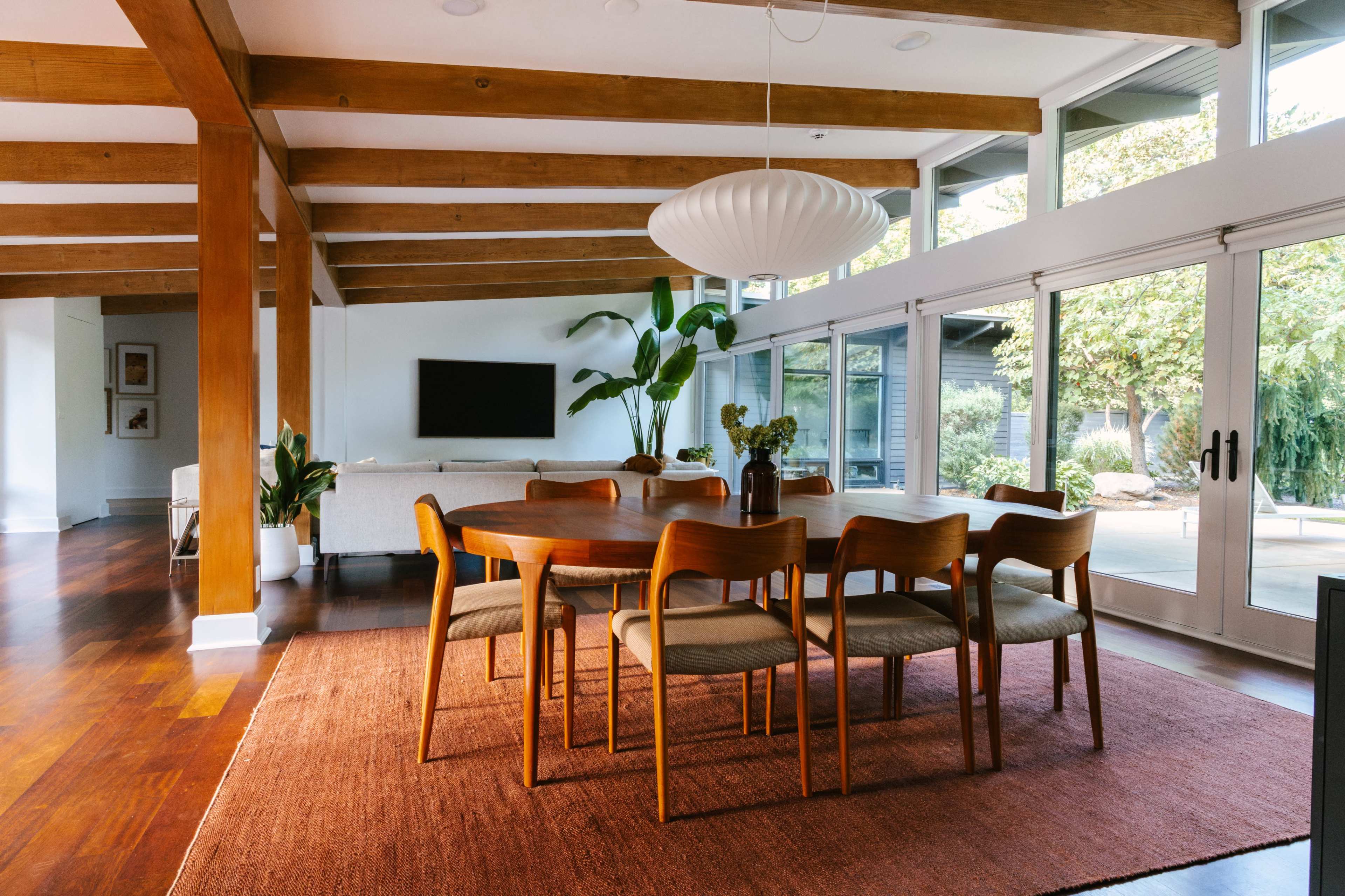 A dining area features a wooden table surrounded by six chairs, with large windows providing natural light and views of plants outside.