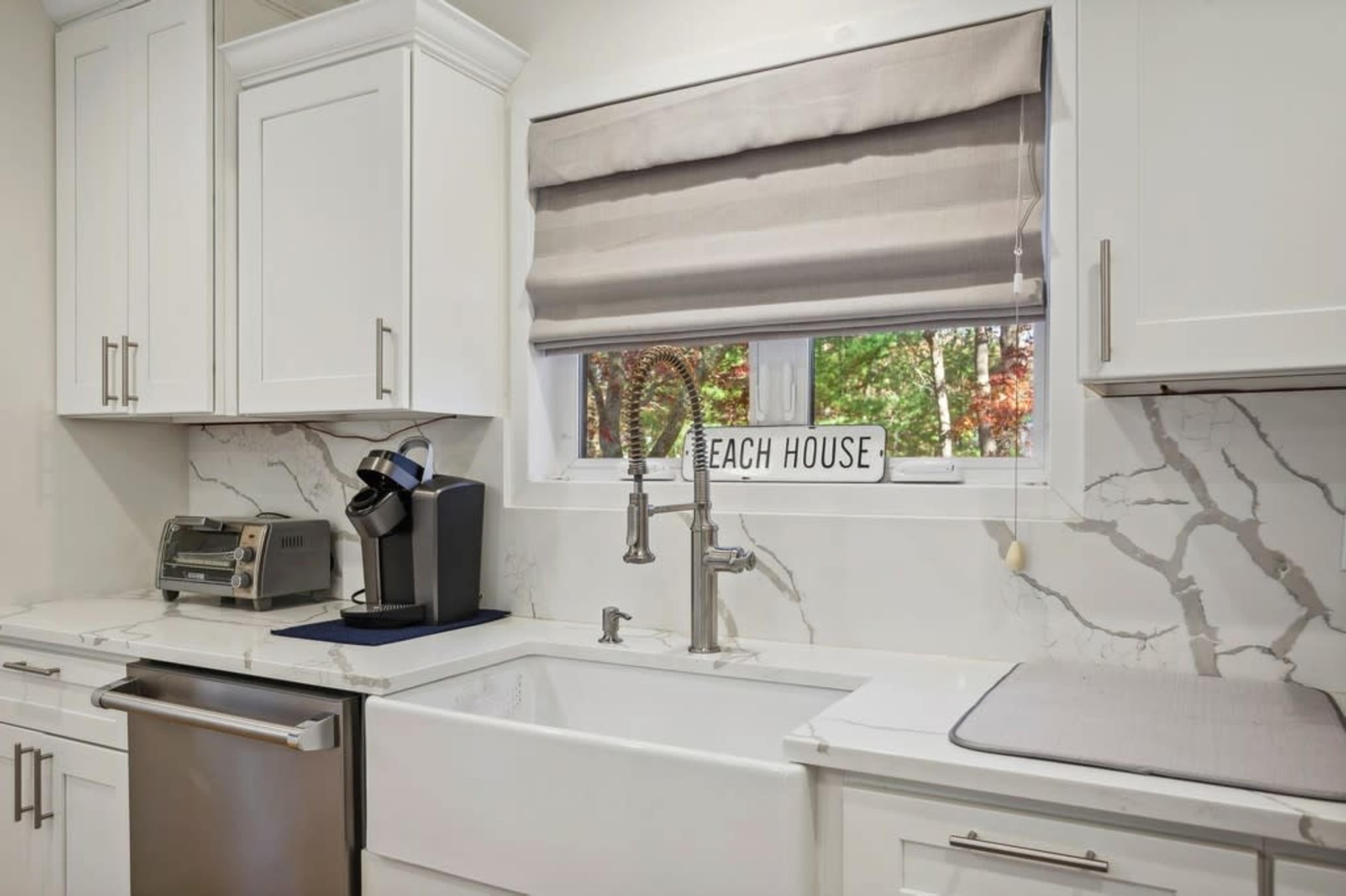 A modern kitchen features white cabinets, a large farmhouse sink, a coffee maker, and a window with gray shades overlooking trees.
