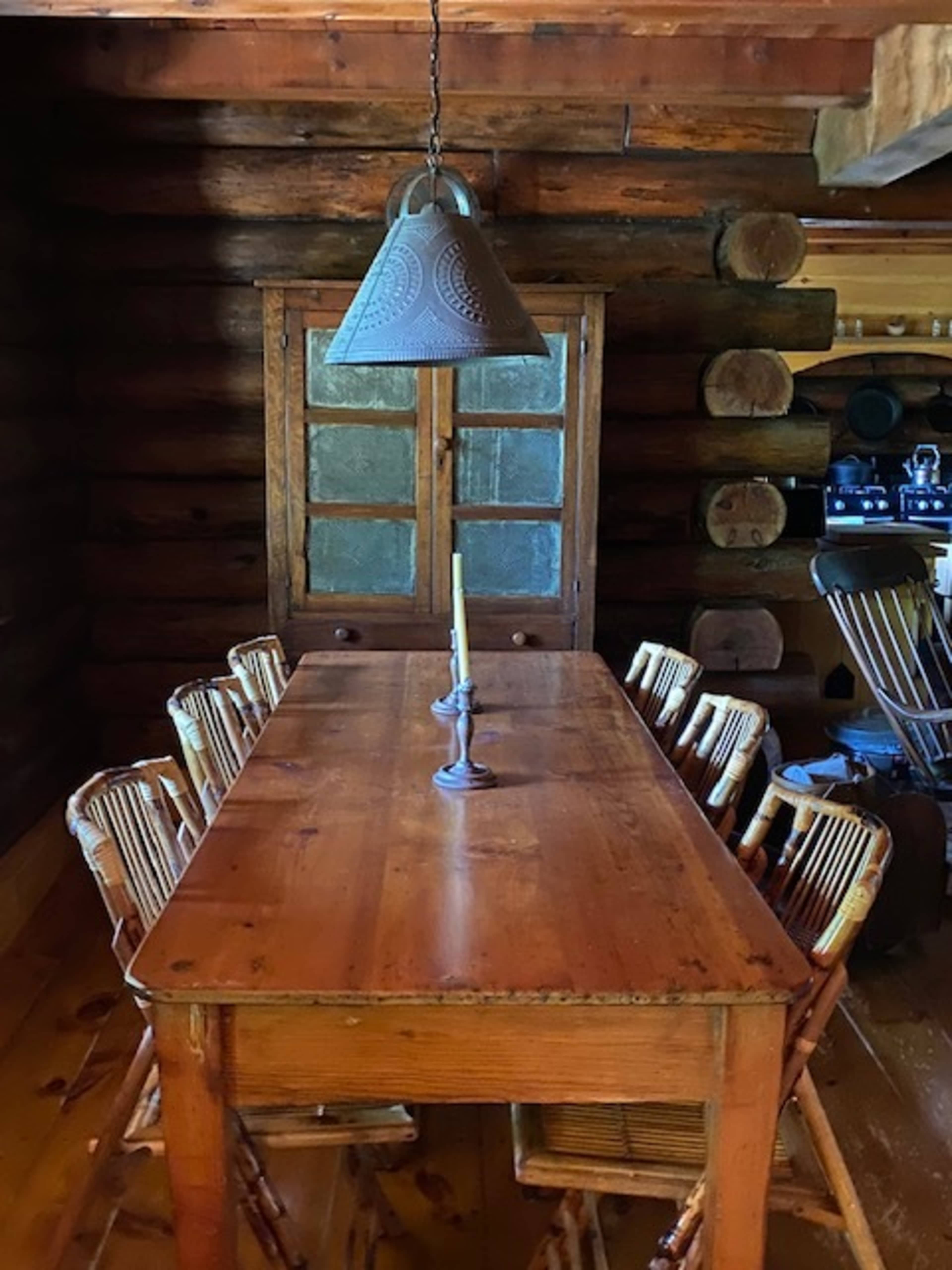 A wooden dining table with eight chairs is set under a hanging light fixture in a rustic log cabin.