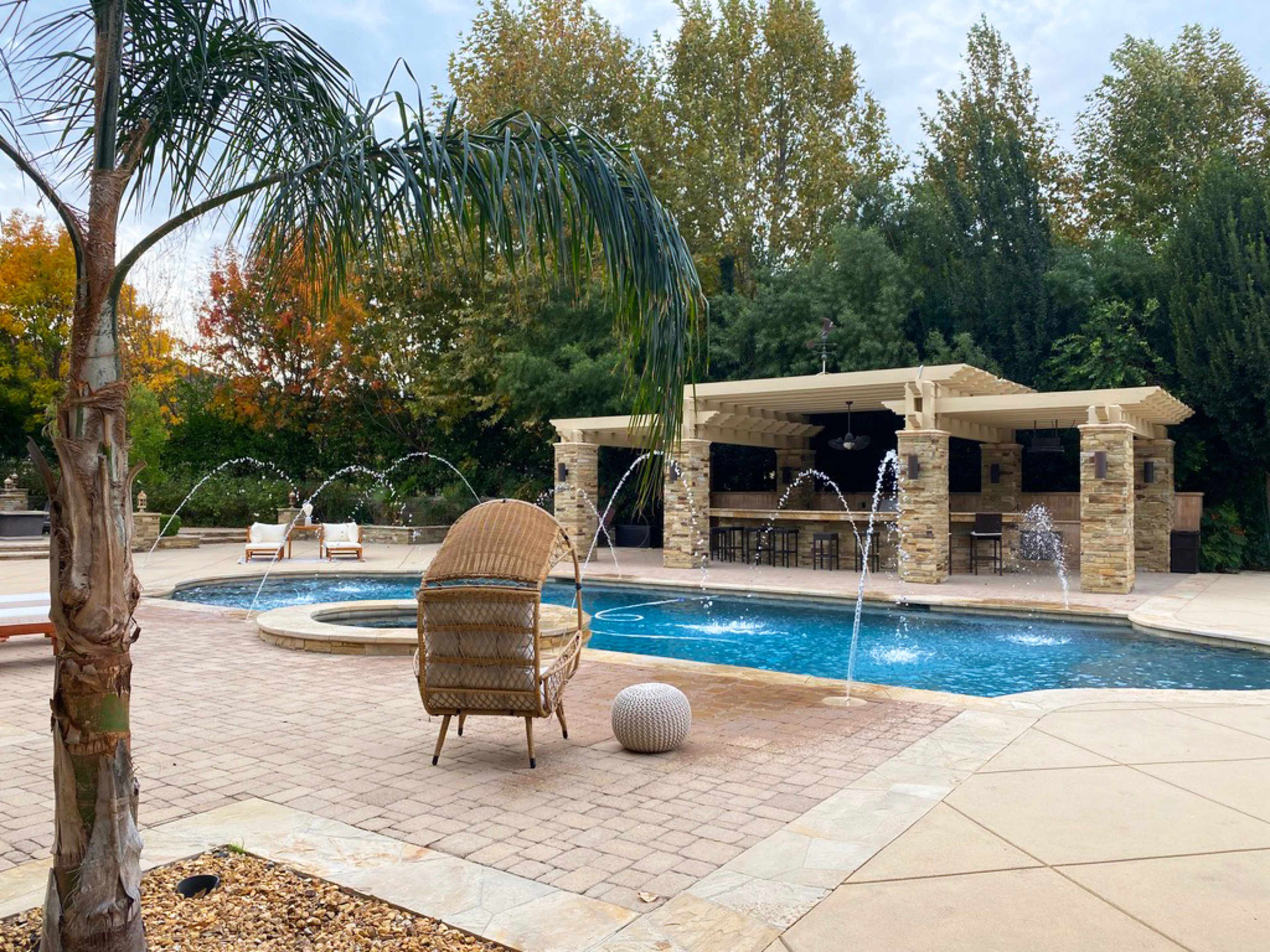 A backyard pool area features a stone patio, a spa, water fountains, and a shaded seating area with a bar.