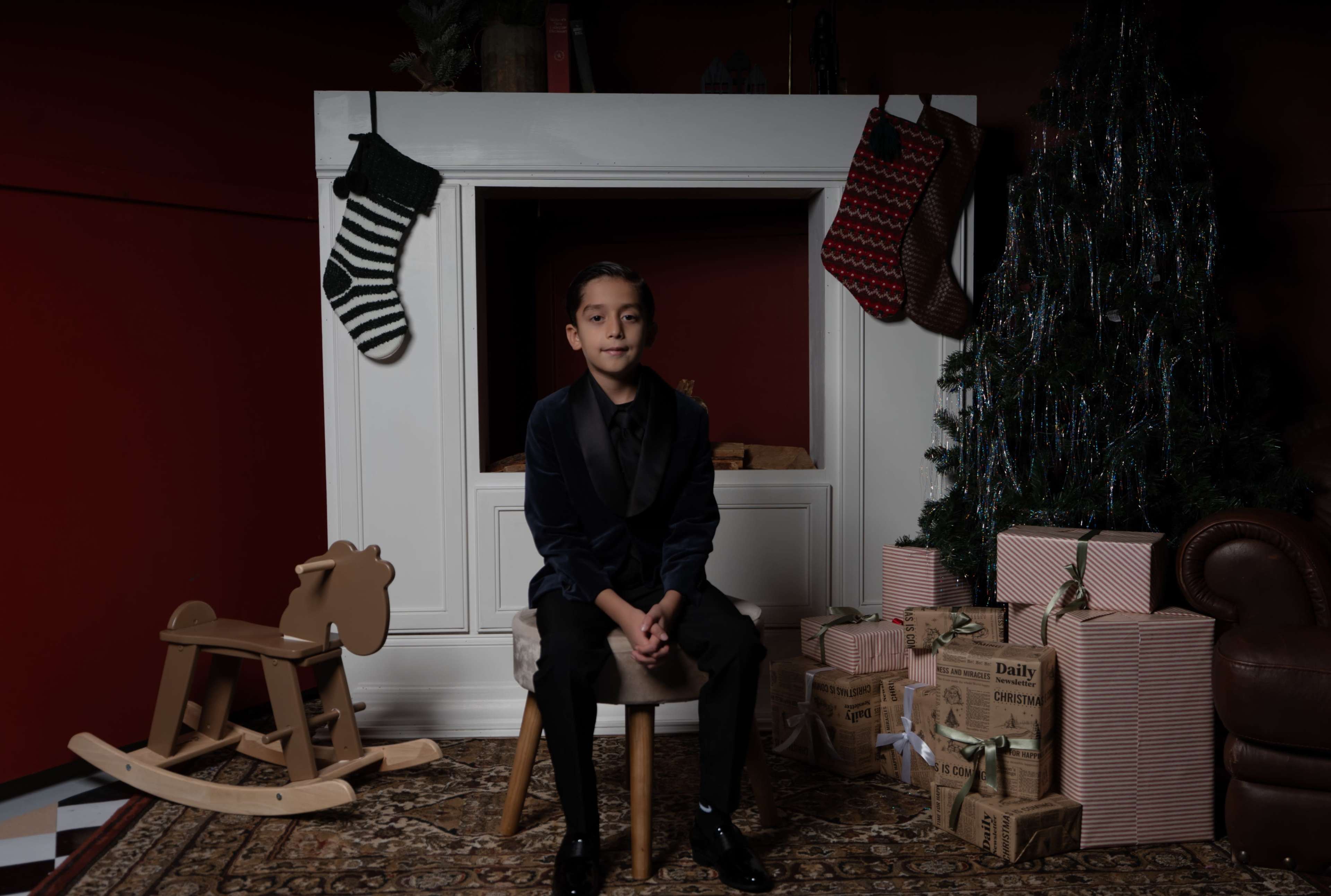 A boy sits on a stool in front of a decorated fireplace with stockings, a Christmas tree, and wrapped presents.