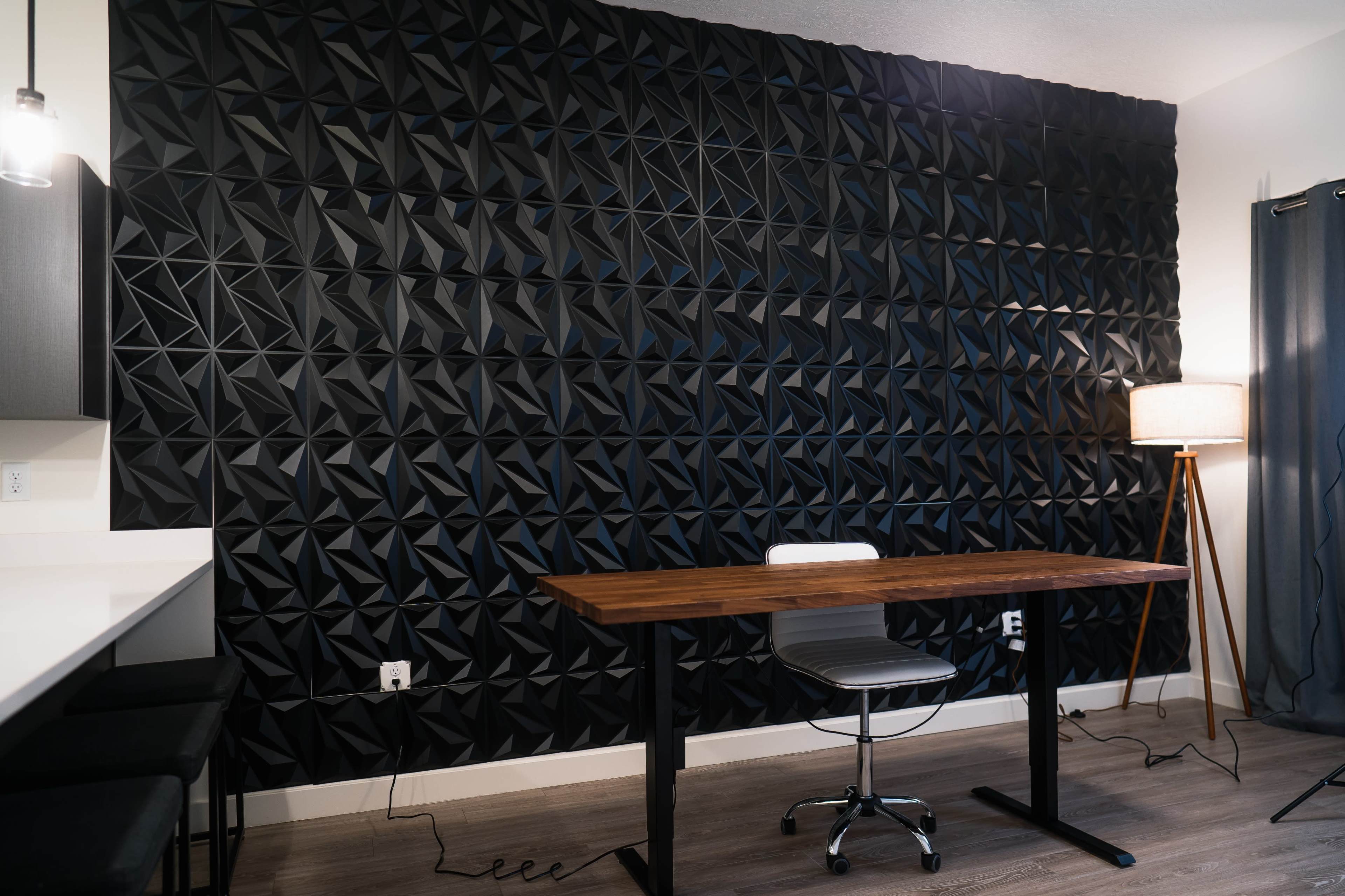 A modern workspace featuring a black geometric wall panel, a wooden desk, a gray chair, and a standing lamp.