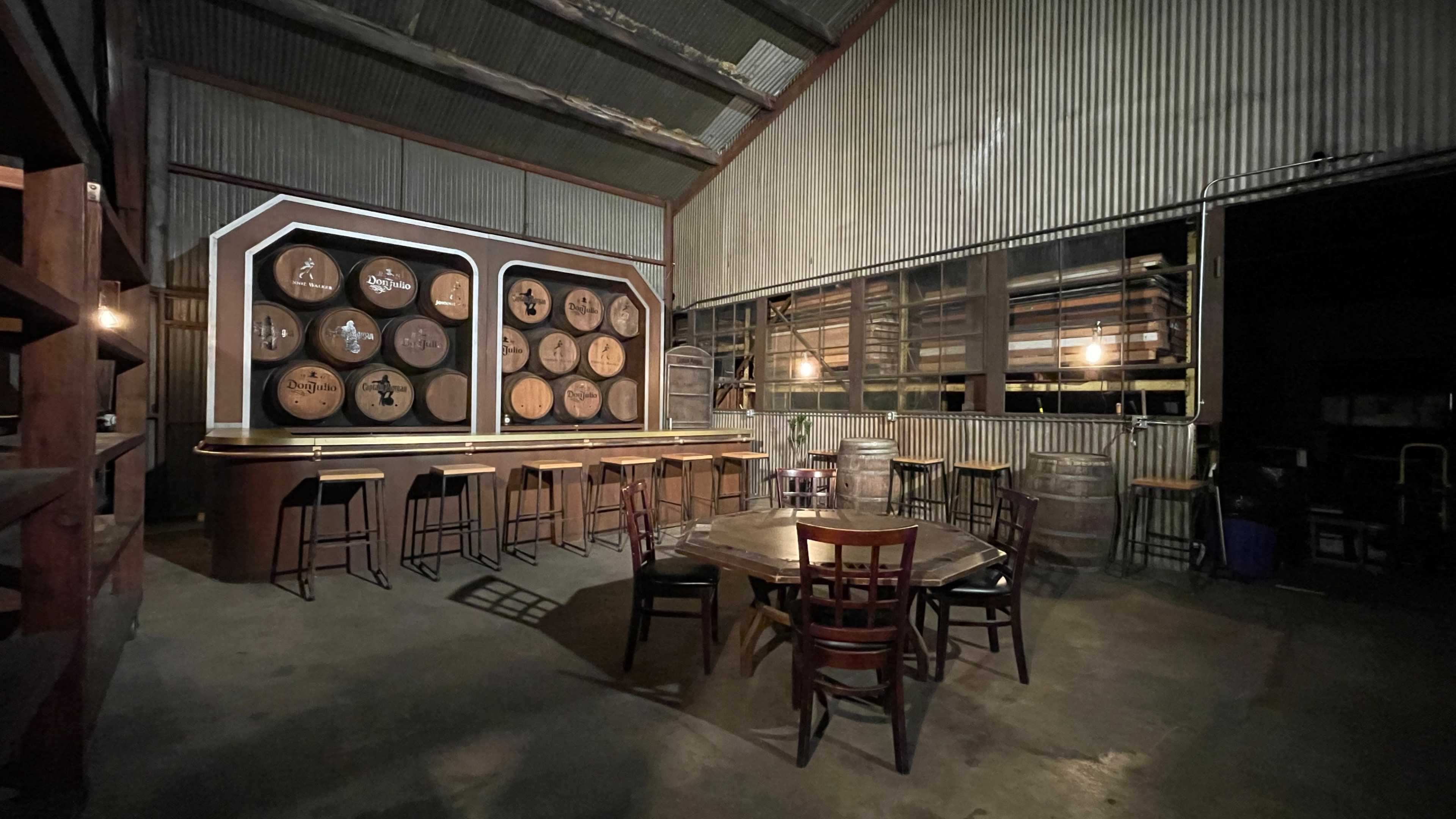 The interior of a rustic warehouse features a bar area with wooden stools, a wall displaying barrels, and a round table surrounded by chairs.