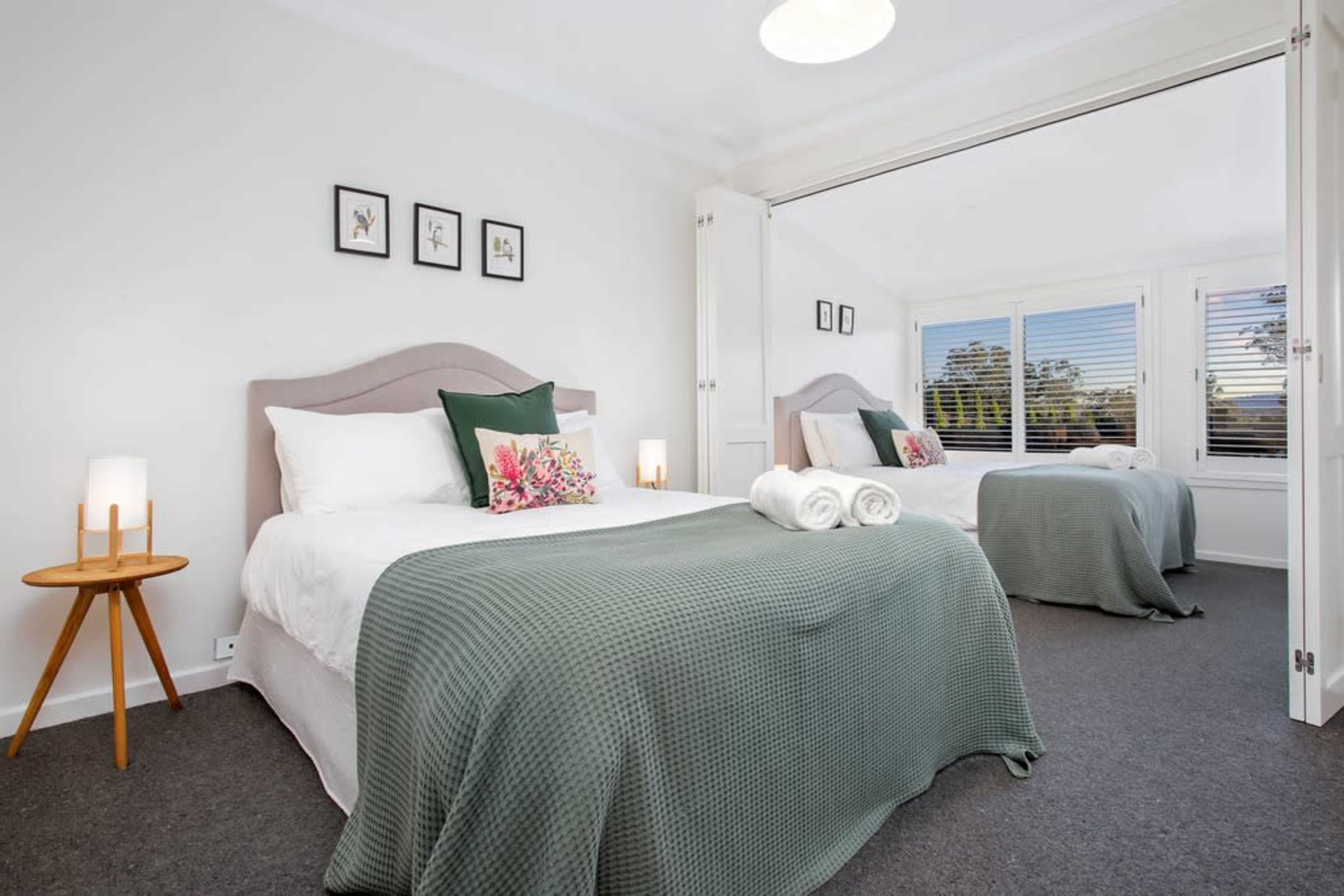 A well-lit bedroom features two beds with white linens and green blankets, accompanied by a bedside table and decorative artwork on the walls.