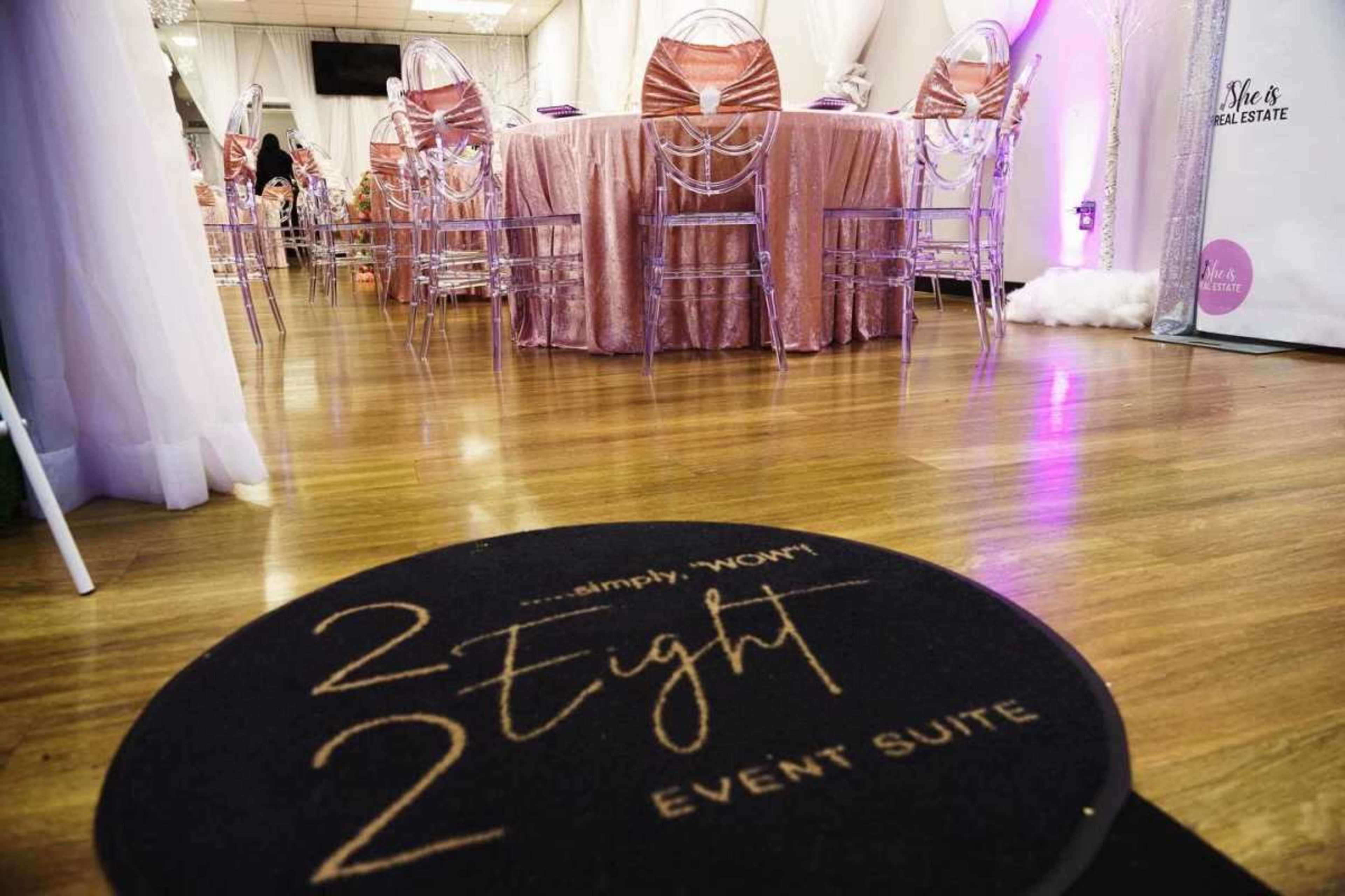 A decorated event space featuring a round black rug with the text "22 Eight Event Suite" and tables set with pink tablecloths and clear chiavari chairs.