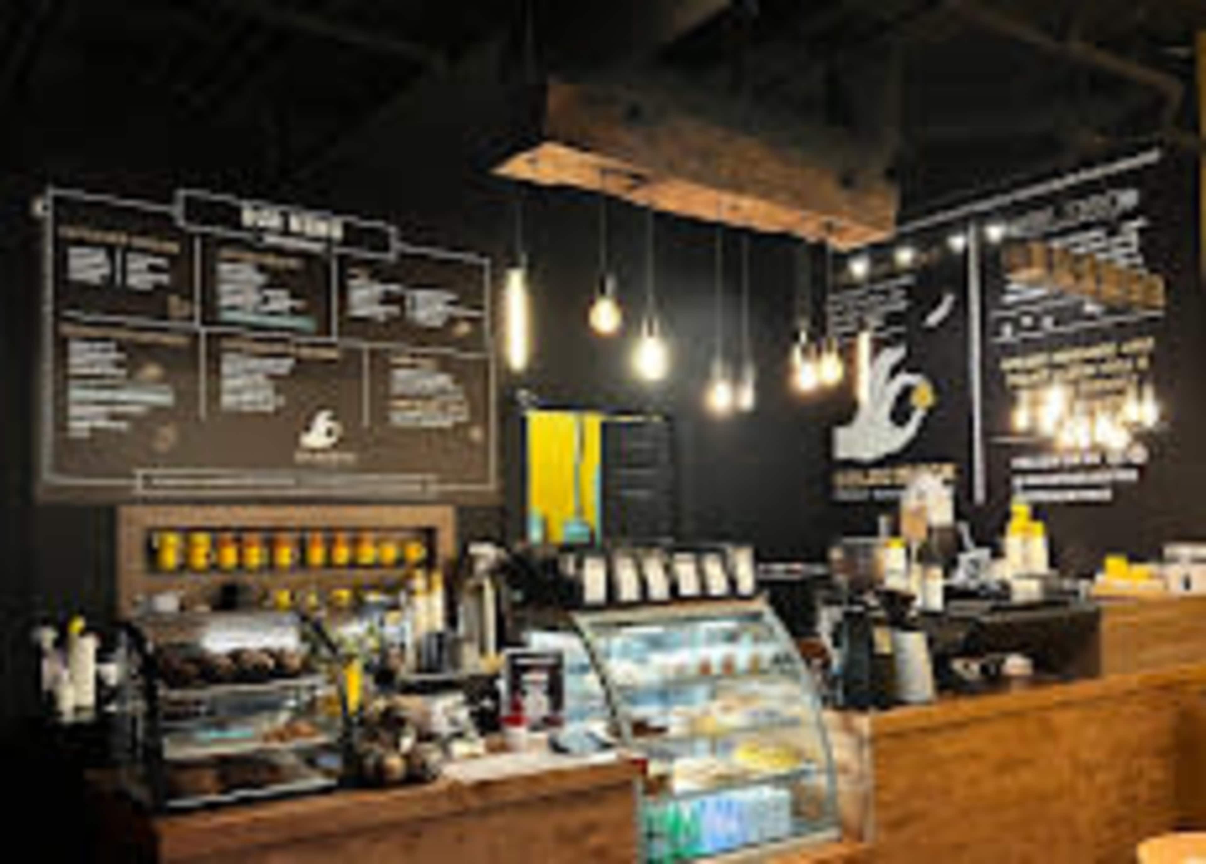 The image shows a coffee shop interior with a wooden counter, display case for food, various menu items on the wall, and hanging light fixtures.