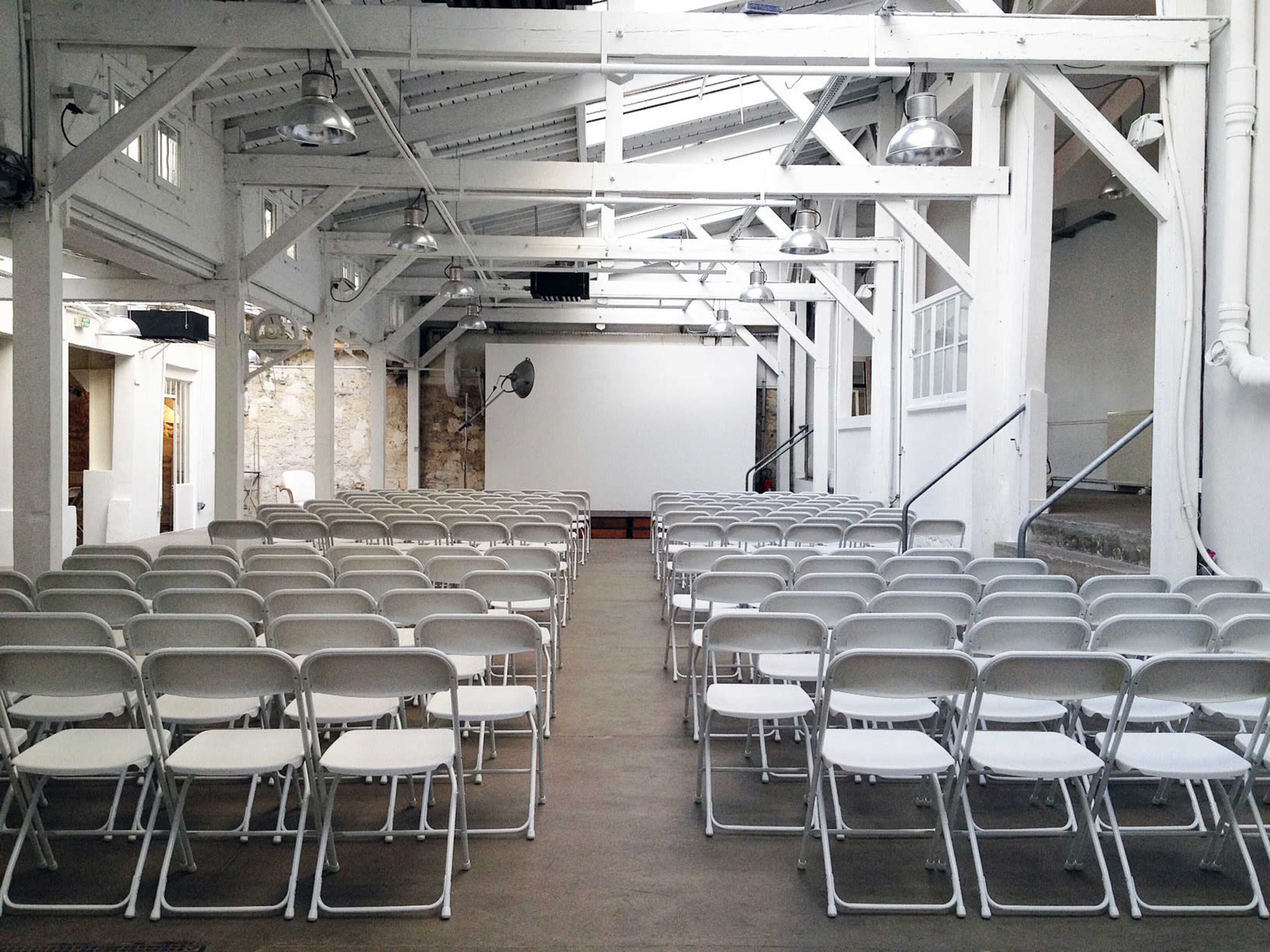 A venue features rows of empty white chairs facing a large projection screen in a bright, industrial space.
