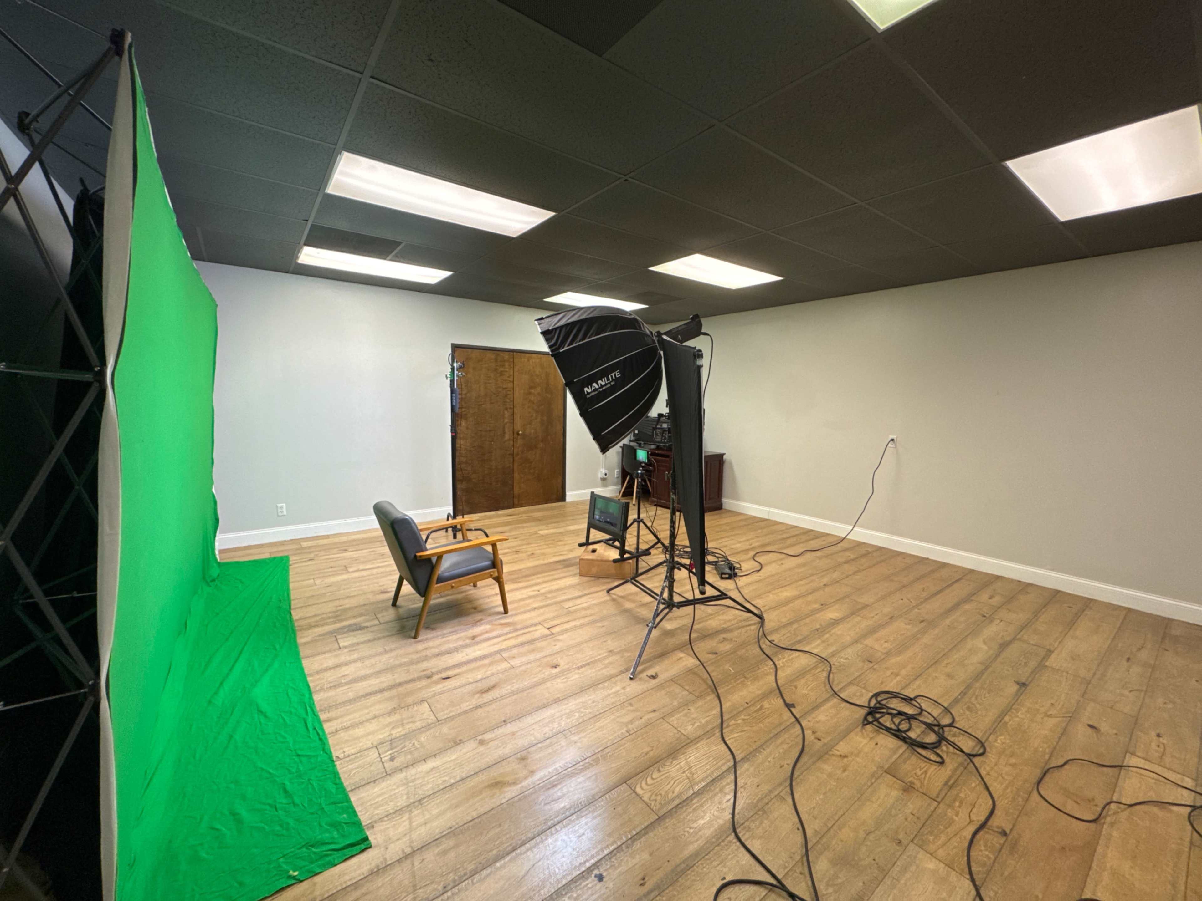 The image shows a photography studio setup with a green backdrop, a chair, and lighting equipment on a wooden floor.