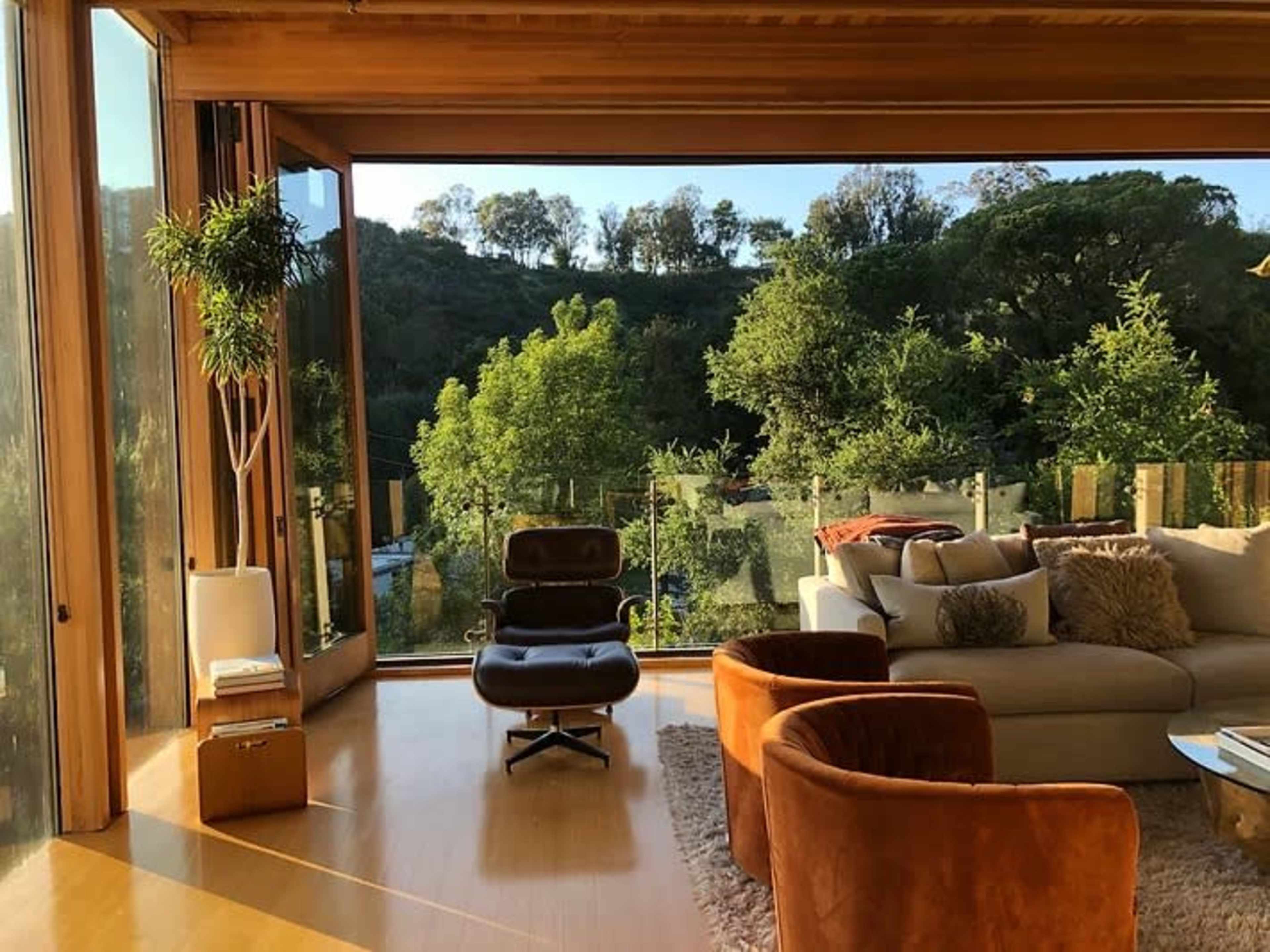 The image shows a modern living room with large windows offering a view of lush greenery and a serene outdoor space.