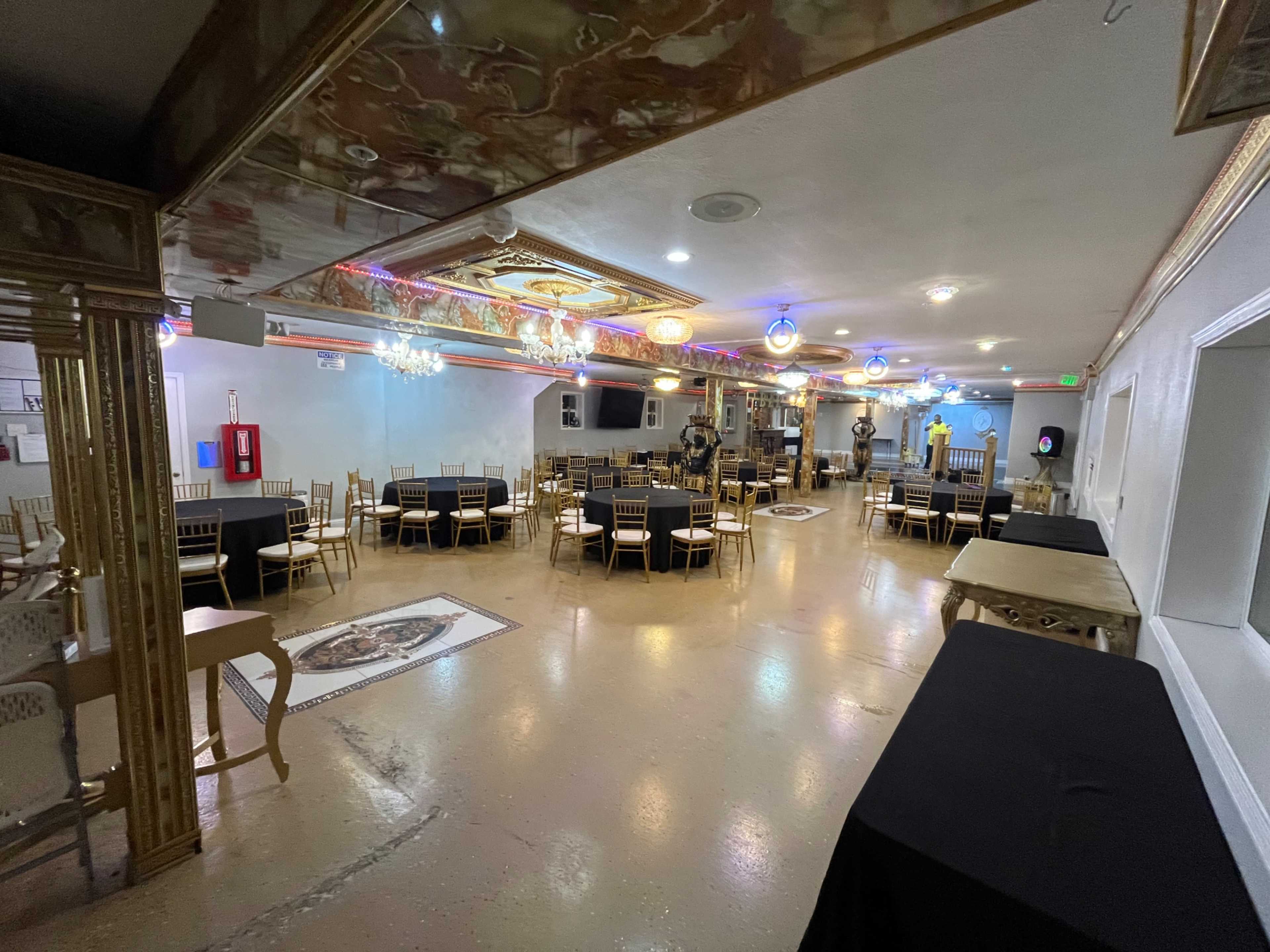 The image shows a spacious event venue featuring neatly arranged tables with black tablecloths, gold accents, and decorative lighting along the ceiling.