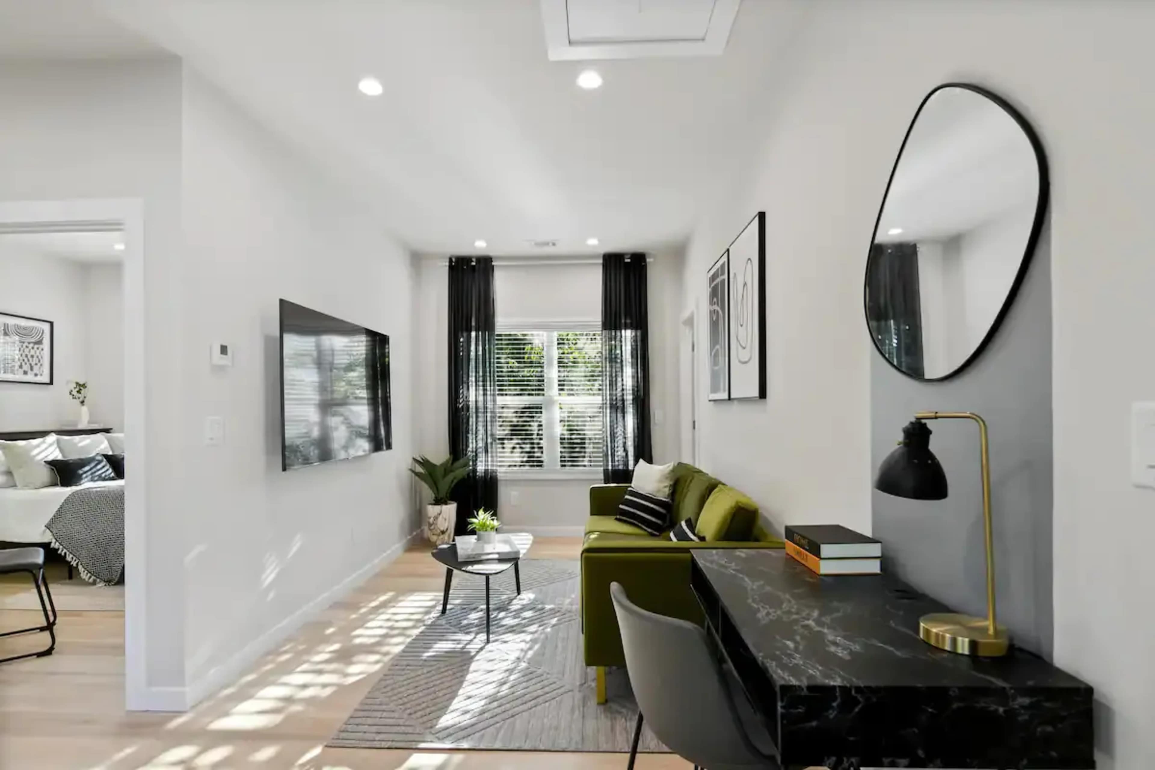 A modern living space featuring a green sofa, a black marble desk with a lamp, and a decorative plant, illuminated by natural light from large windows.