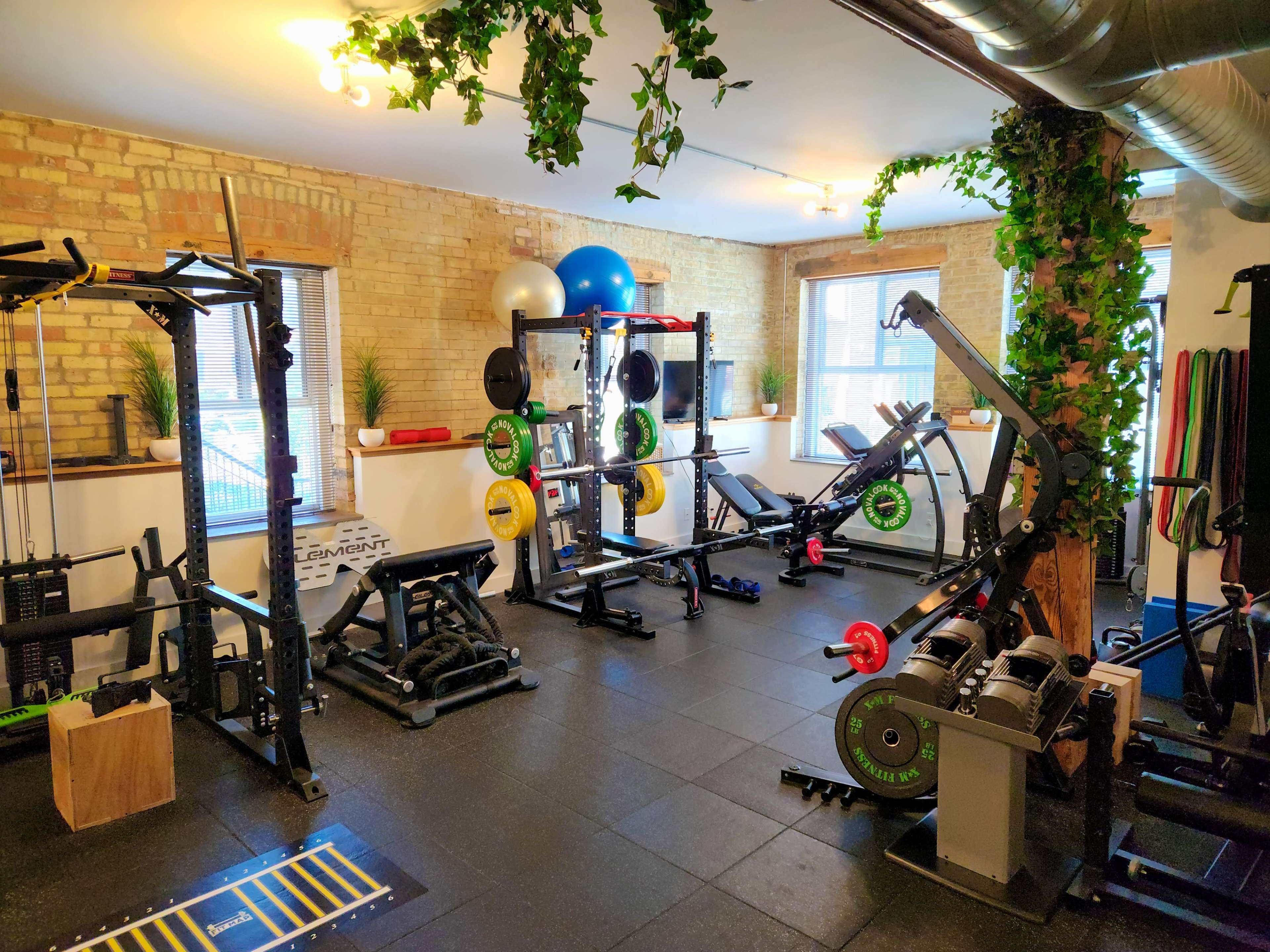 The image shows a well-equipped gym with various weightlifting machines, free weights, exercise balls, and a rubber flooring surface.