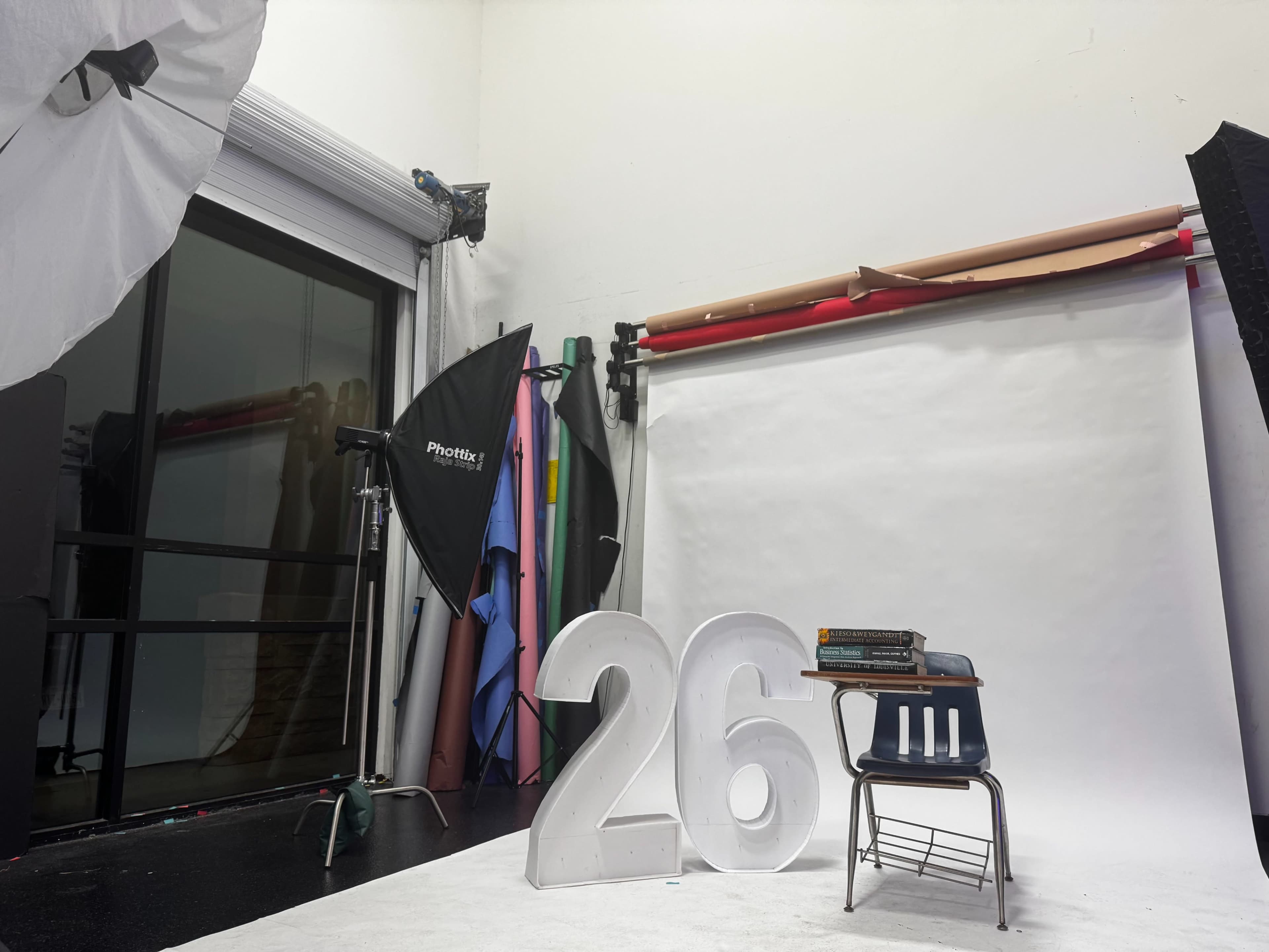 The scene features a photography studio with a large decorative number "26," a wooden chair, and various backdrops and lighting equipment.