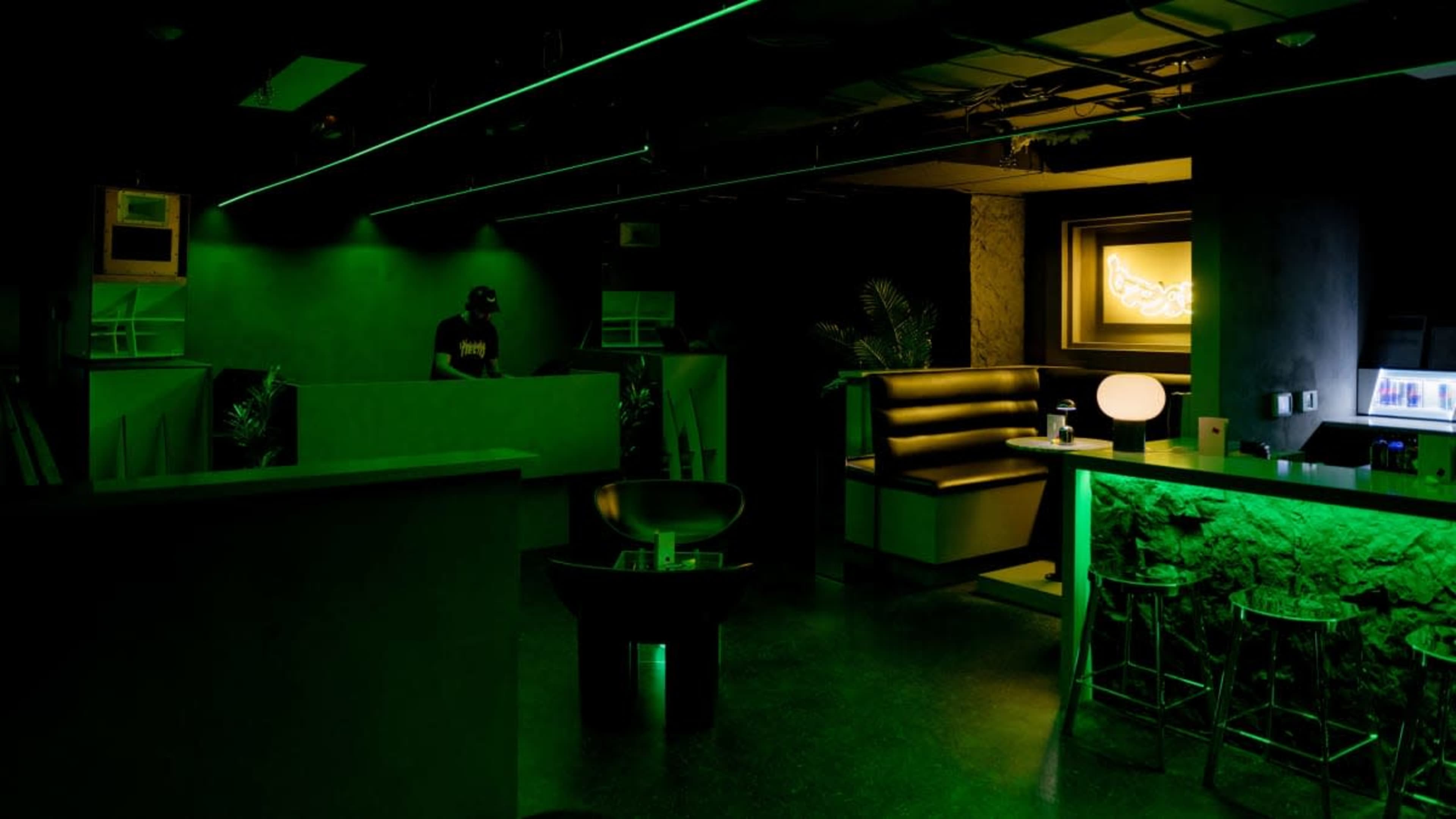 The image shows a dimly lit lounge or bar with green lighting, featuring a DJ booth, seating areas, and illuminated bar counter elements.