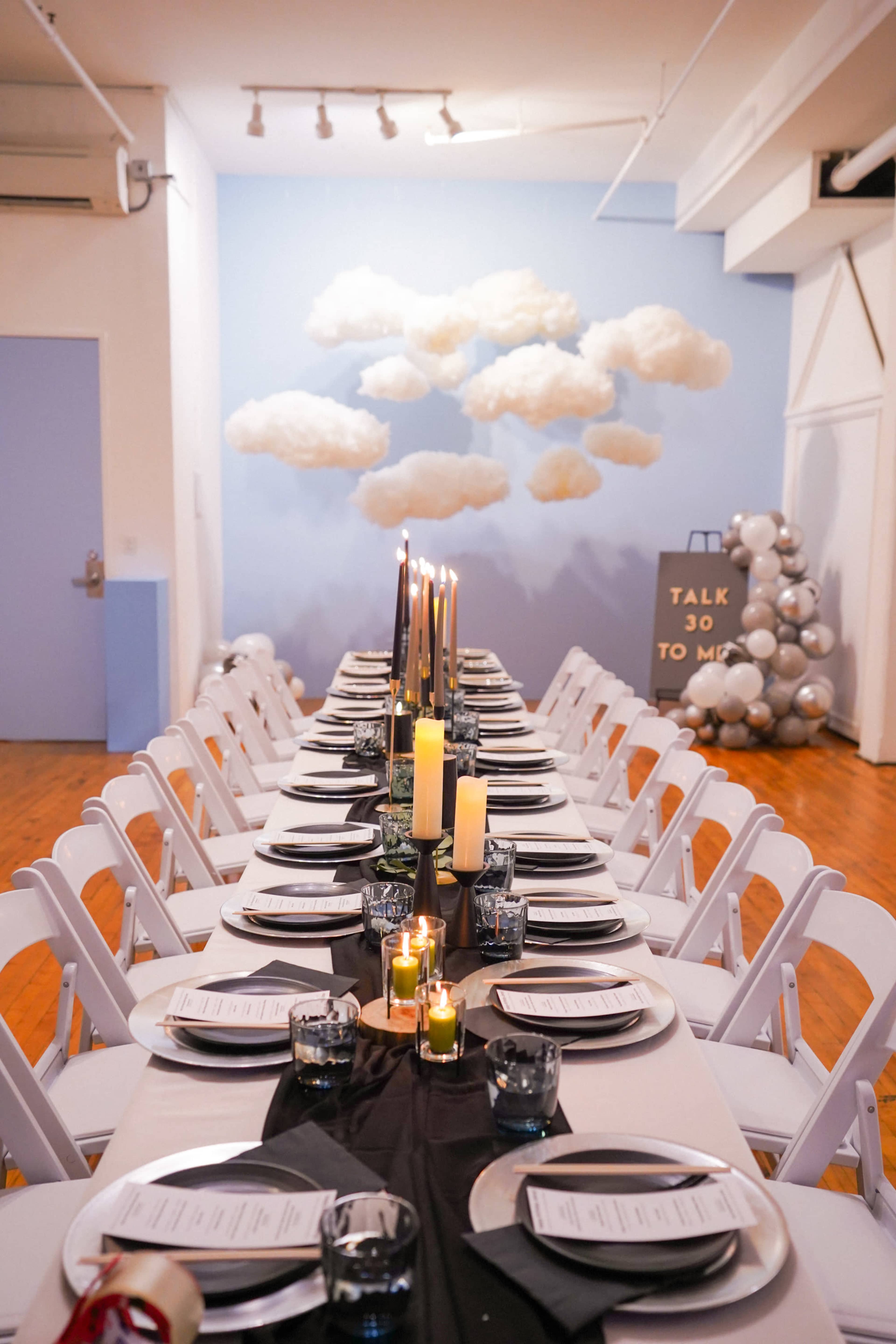 A long dining table is set with candles and decorative plates in a room featuring a cloud-themed backdrop and a sign that reads "TALK 30 TO ME."