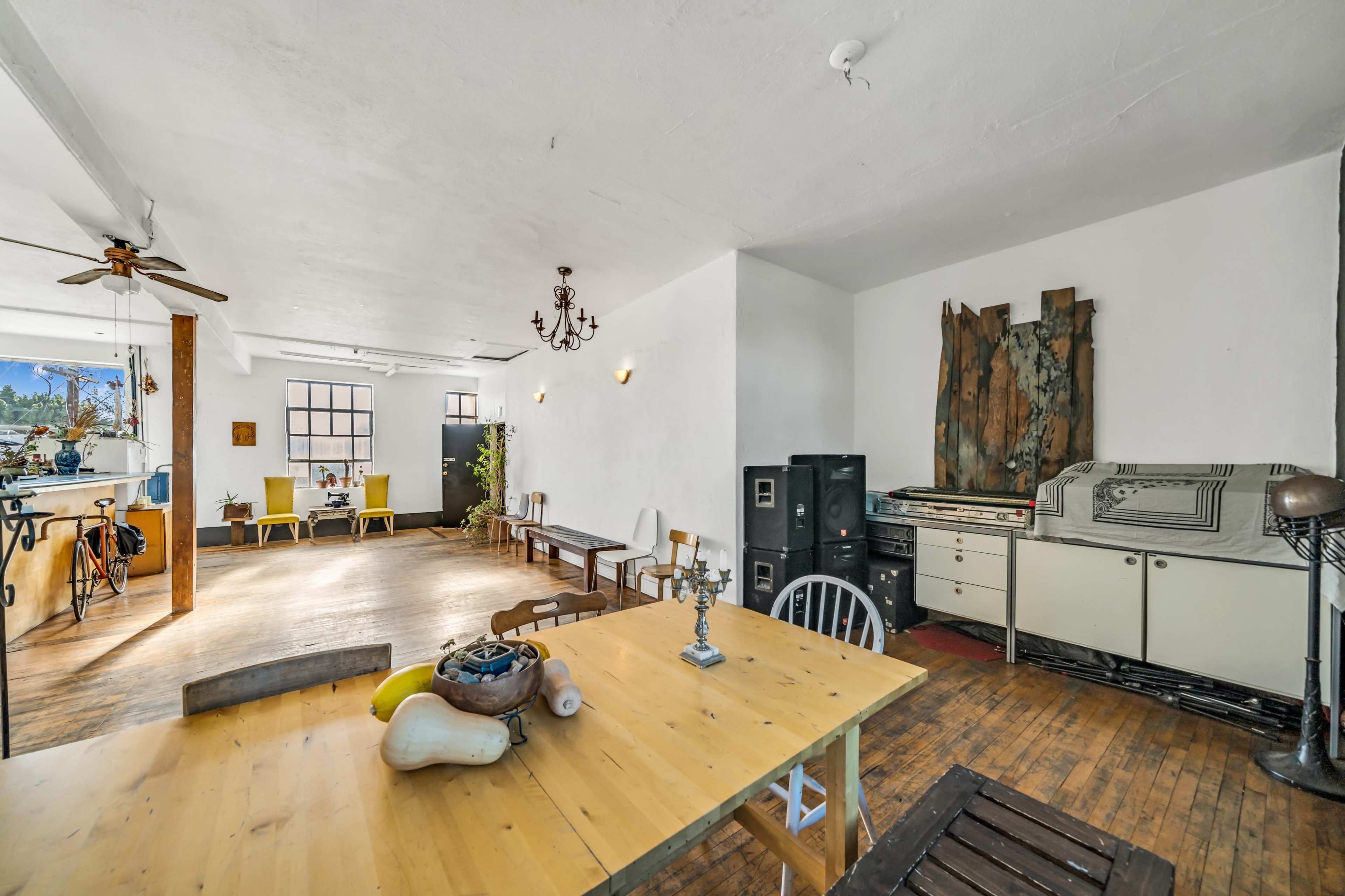 The image depicts a spacious, airy room featuring a dining table with chairs, a vintage dresser, and an open kitchen area.