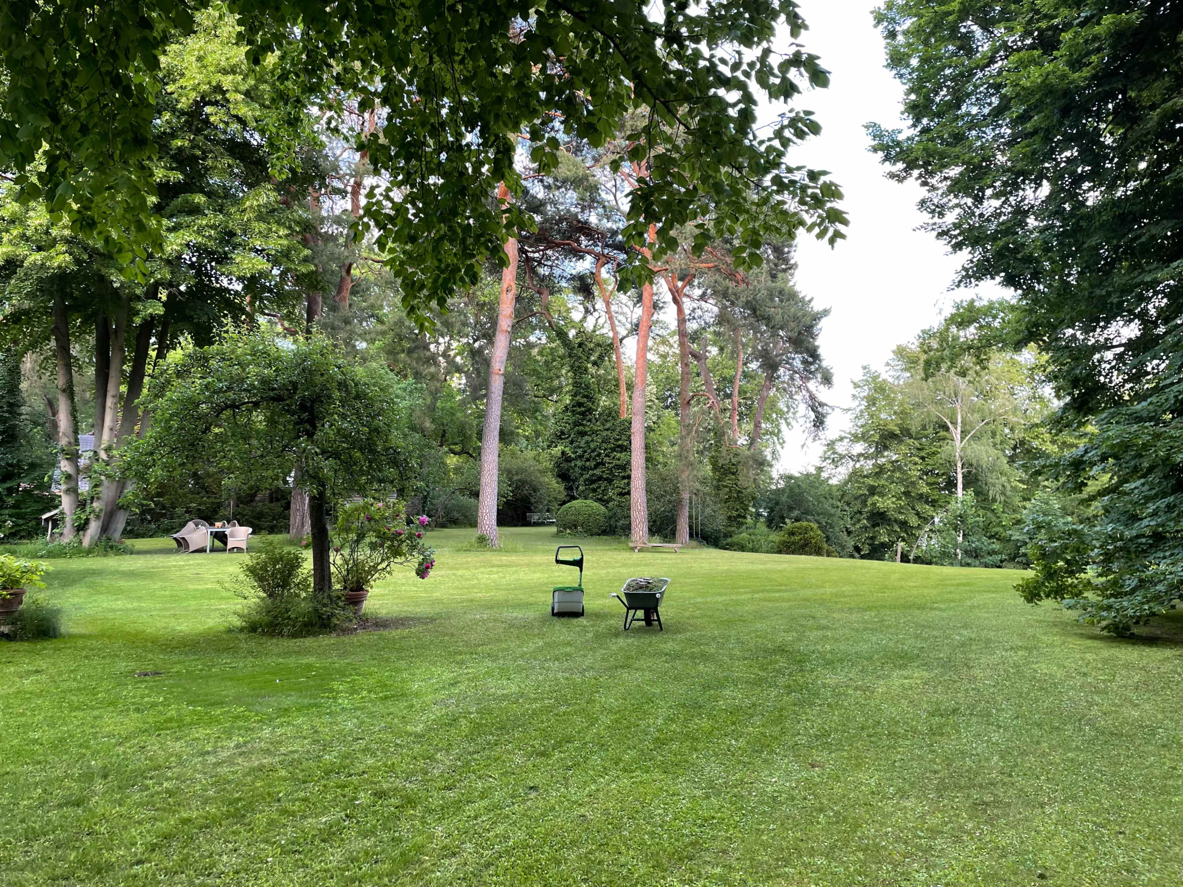 The image shows a lush, green garden with neatly mowed grass, a wheelbarrow, and a garden cart resting on the lawn under tall trees.