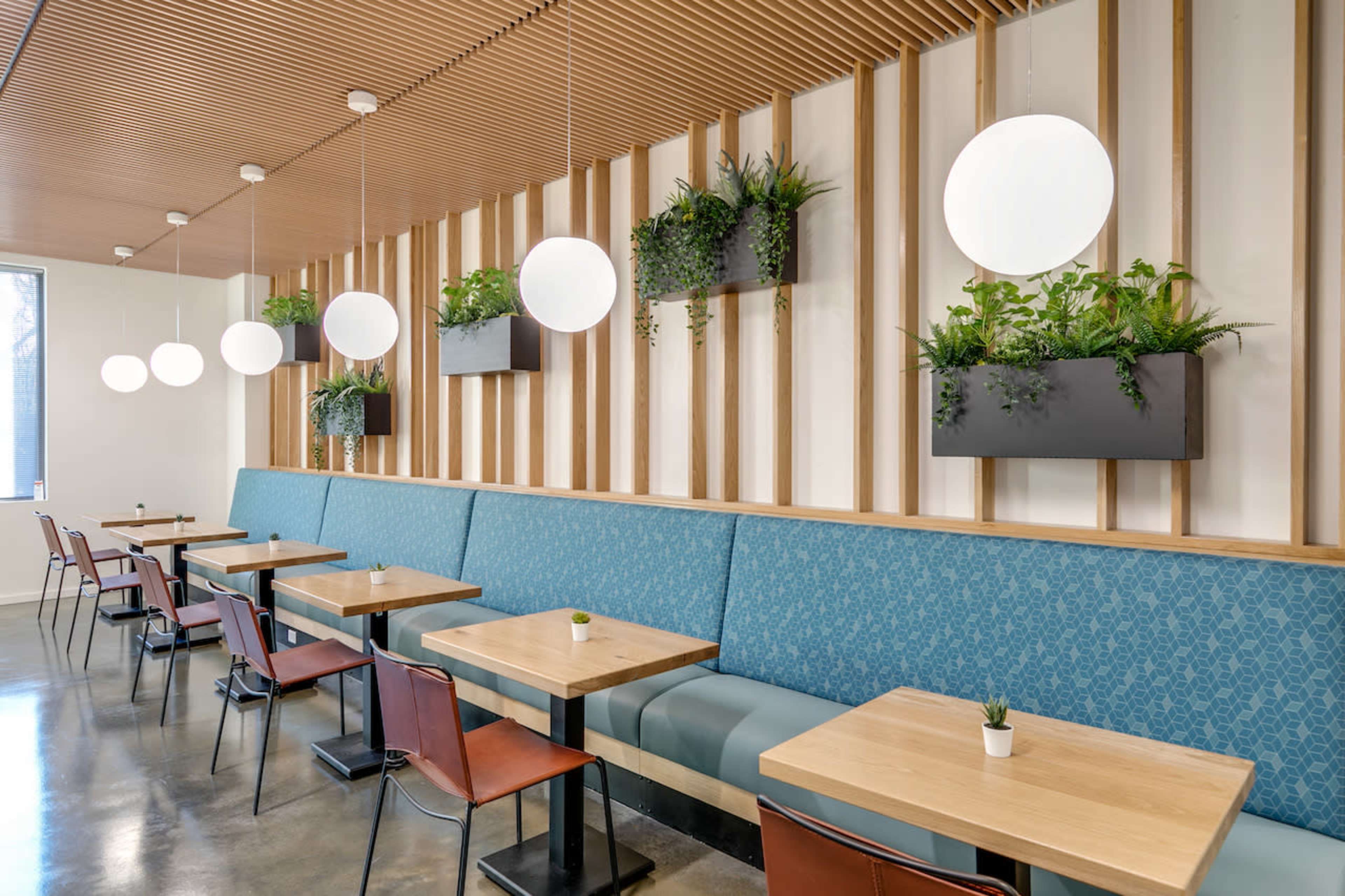 A modern restaurant interior with blue upholstered seating, wooden tables, and round pendant lights hanging above, complemented by green plants in wall planters.