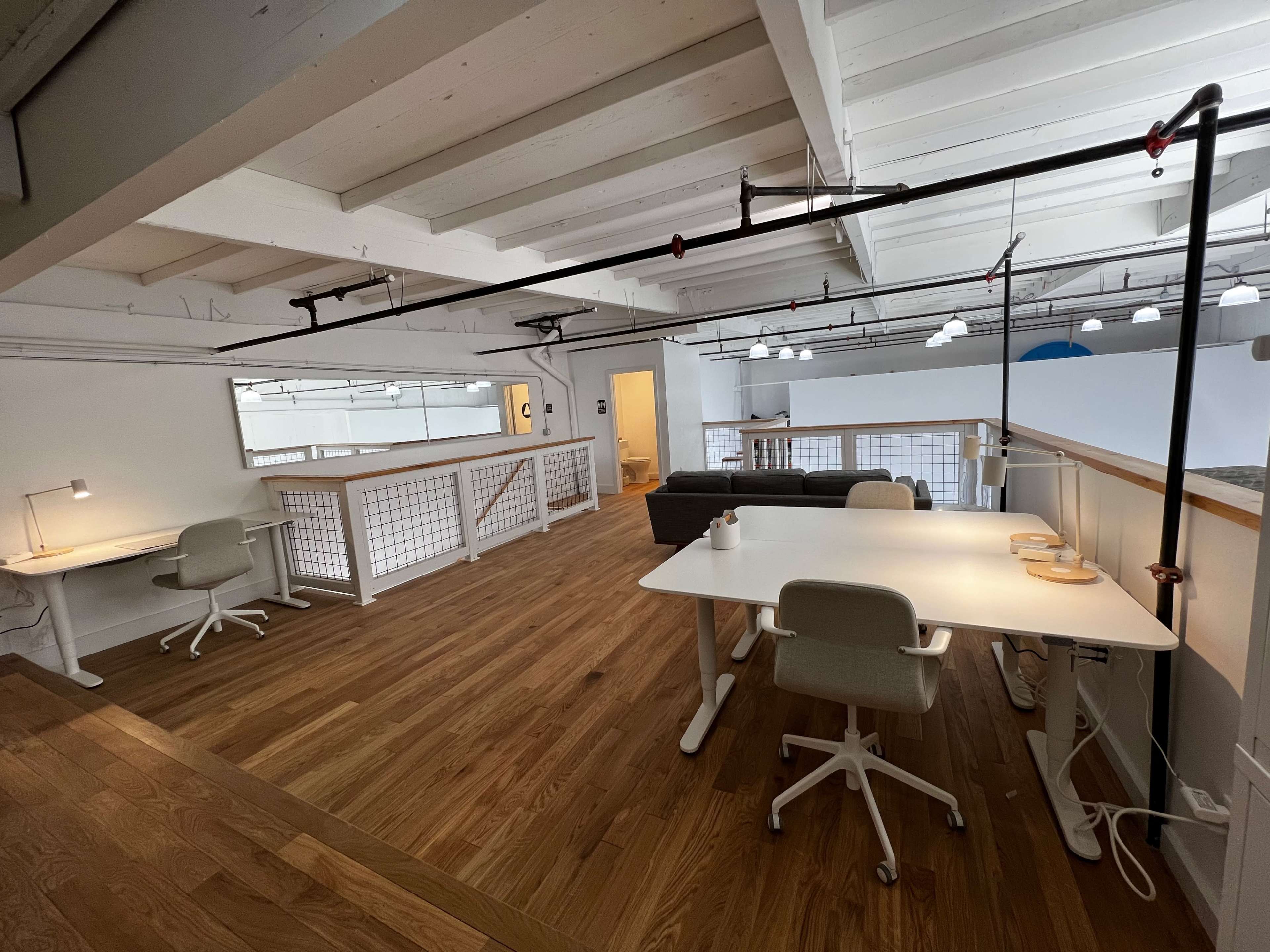 A spacious, modern office interior with wooden flooring, two workstations, a sofa, and overhead lighting.