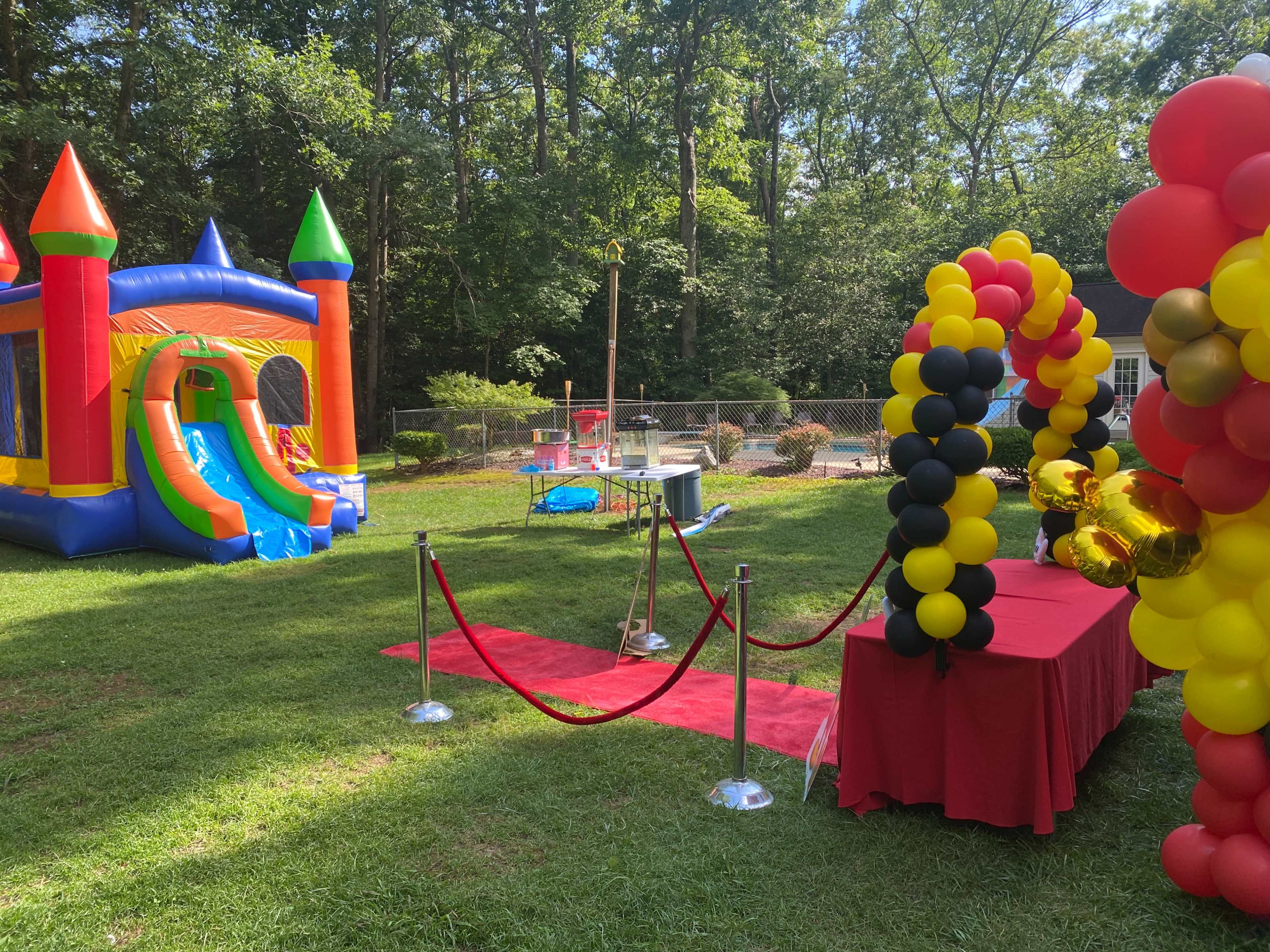 The scene features a colorful bounce house, a red carpet with stanchions, a table draped in red fabric, and a background of greenery and playground equipment in a backyard.
