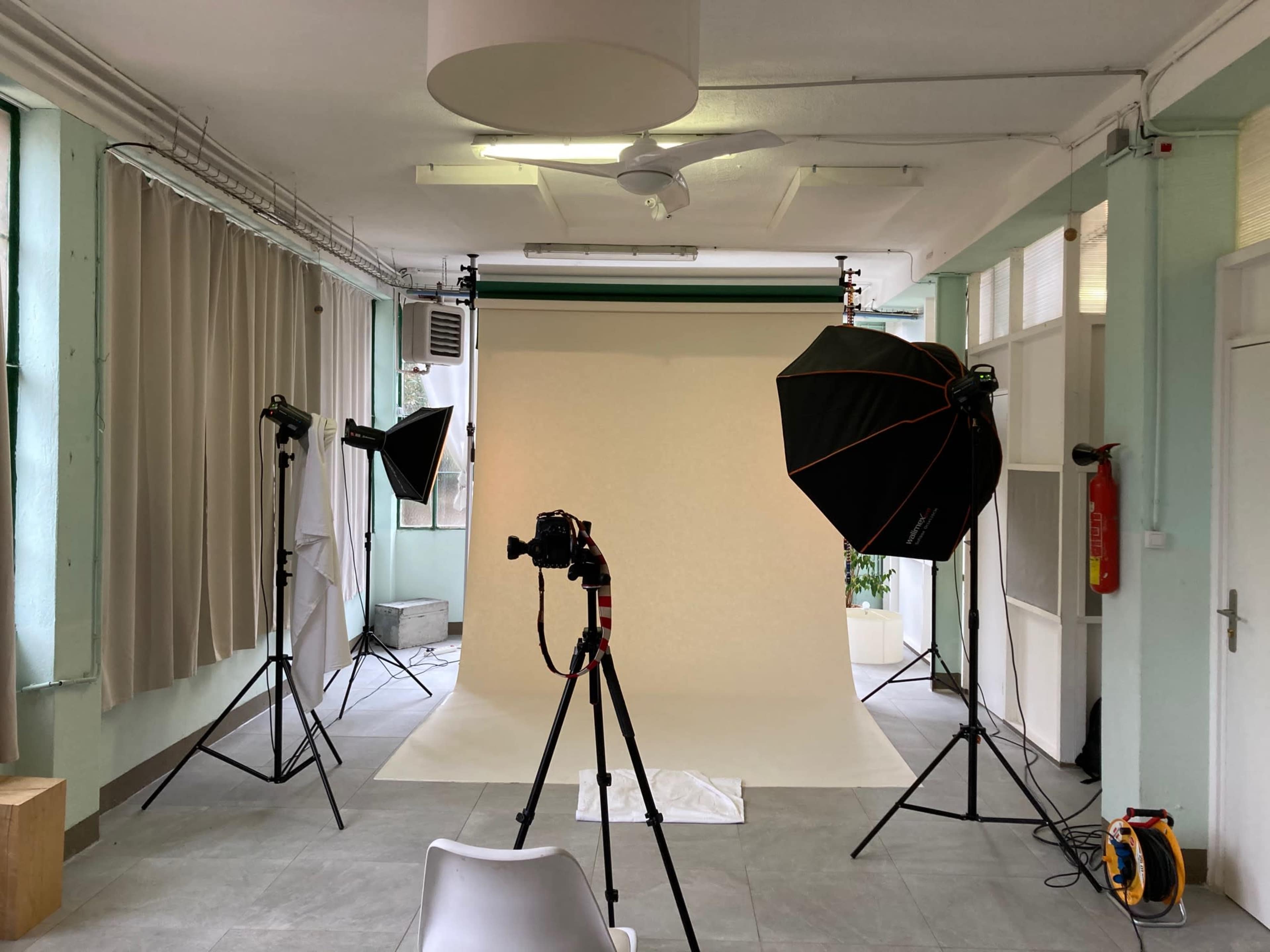 The image shows a photography studio with two light stands, a backdrop, a camera on a tripod, and soft light setups in a spacious, well-lit room.