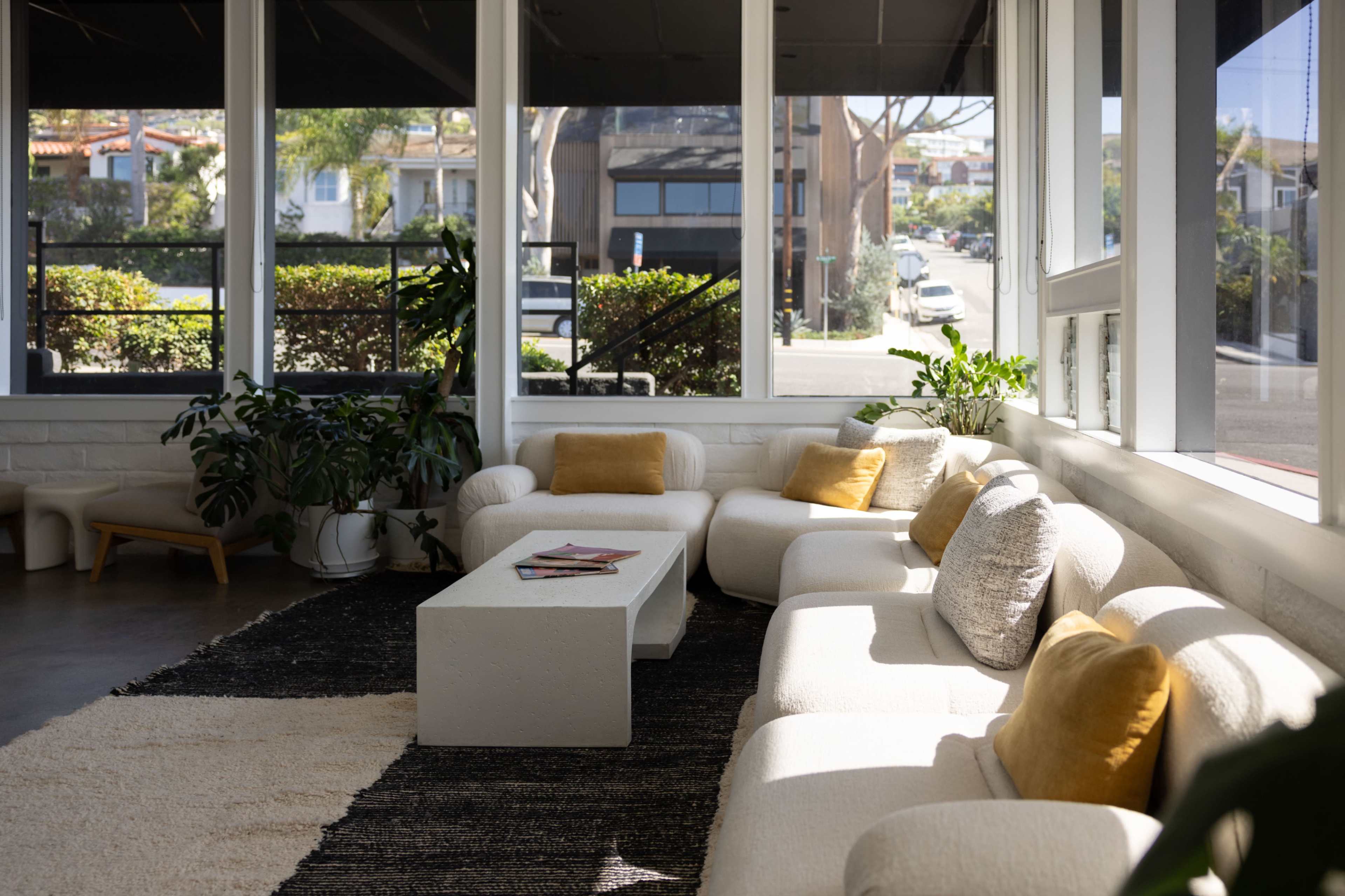 The image shows a bright, modern living space with a white sectional sofa, a coffee table, and plants, framed by large windows.