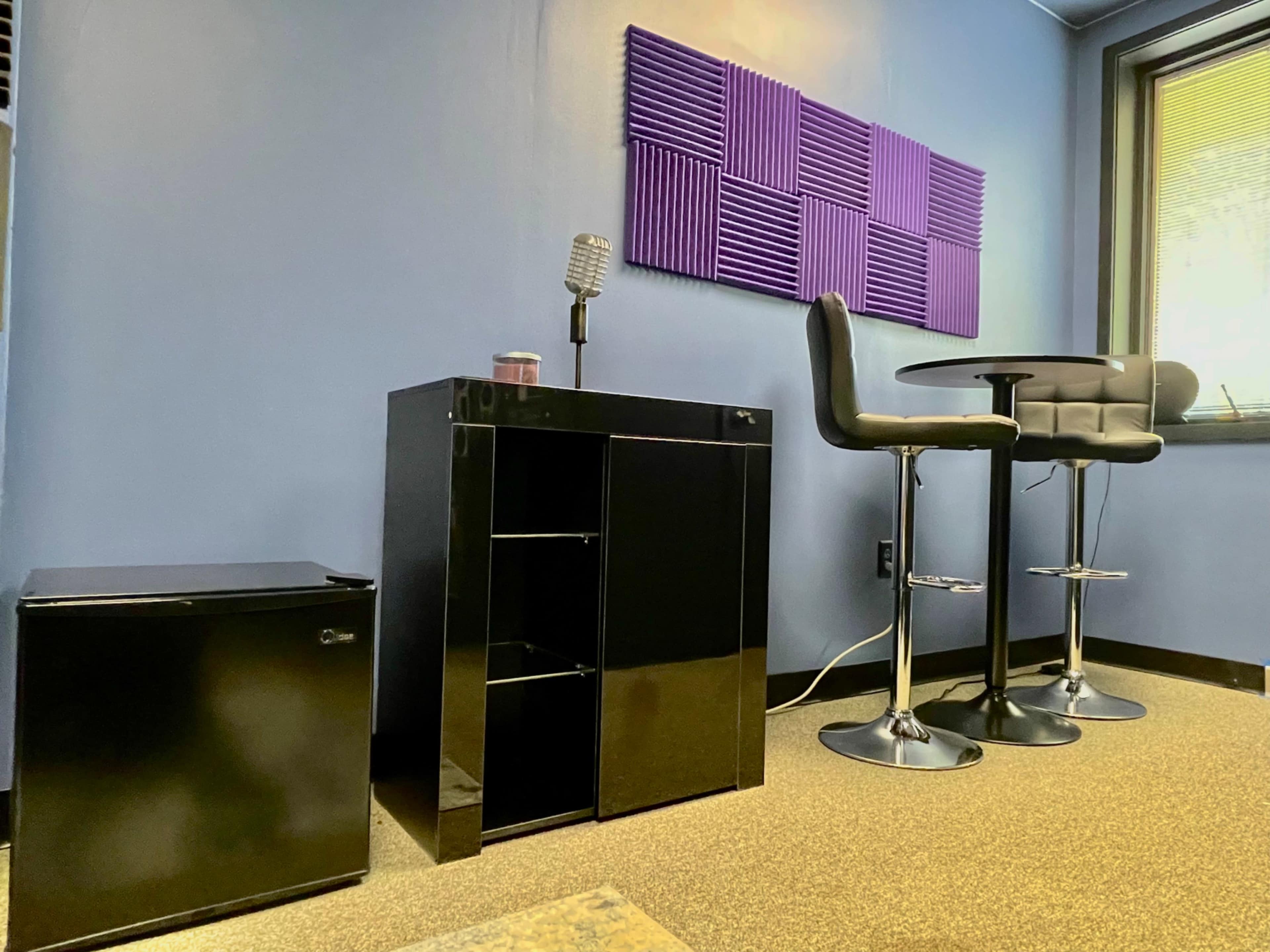 A small office space featuring a black mini refrigerator, a black cabinet, and a round table with two black stools, all against a blue wall with purple soundproofing panels.
