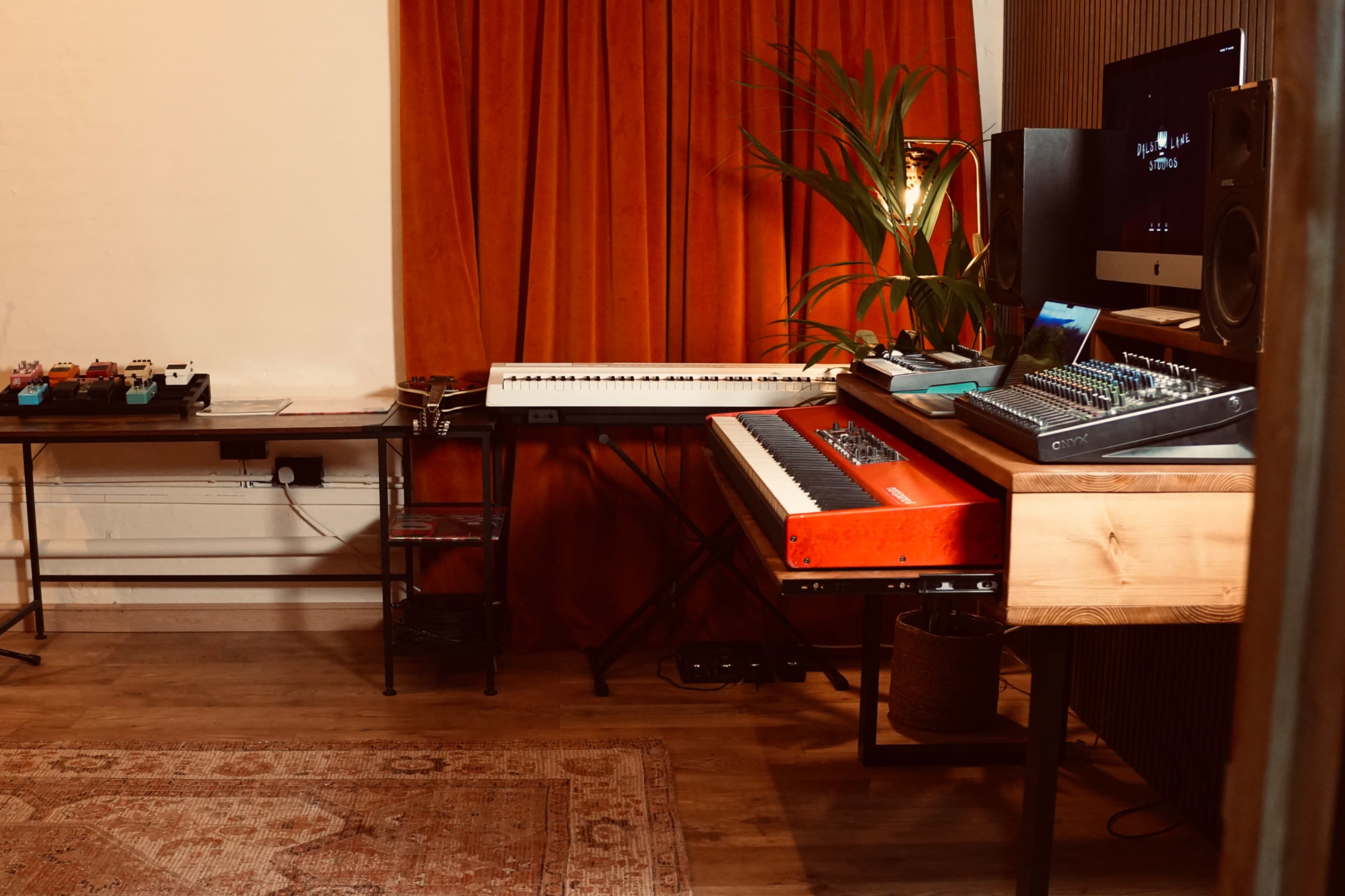 The image shows a music production setup featuring a keyboard, a mixing console, and various audio equipment against a backdrop of red curtains.