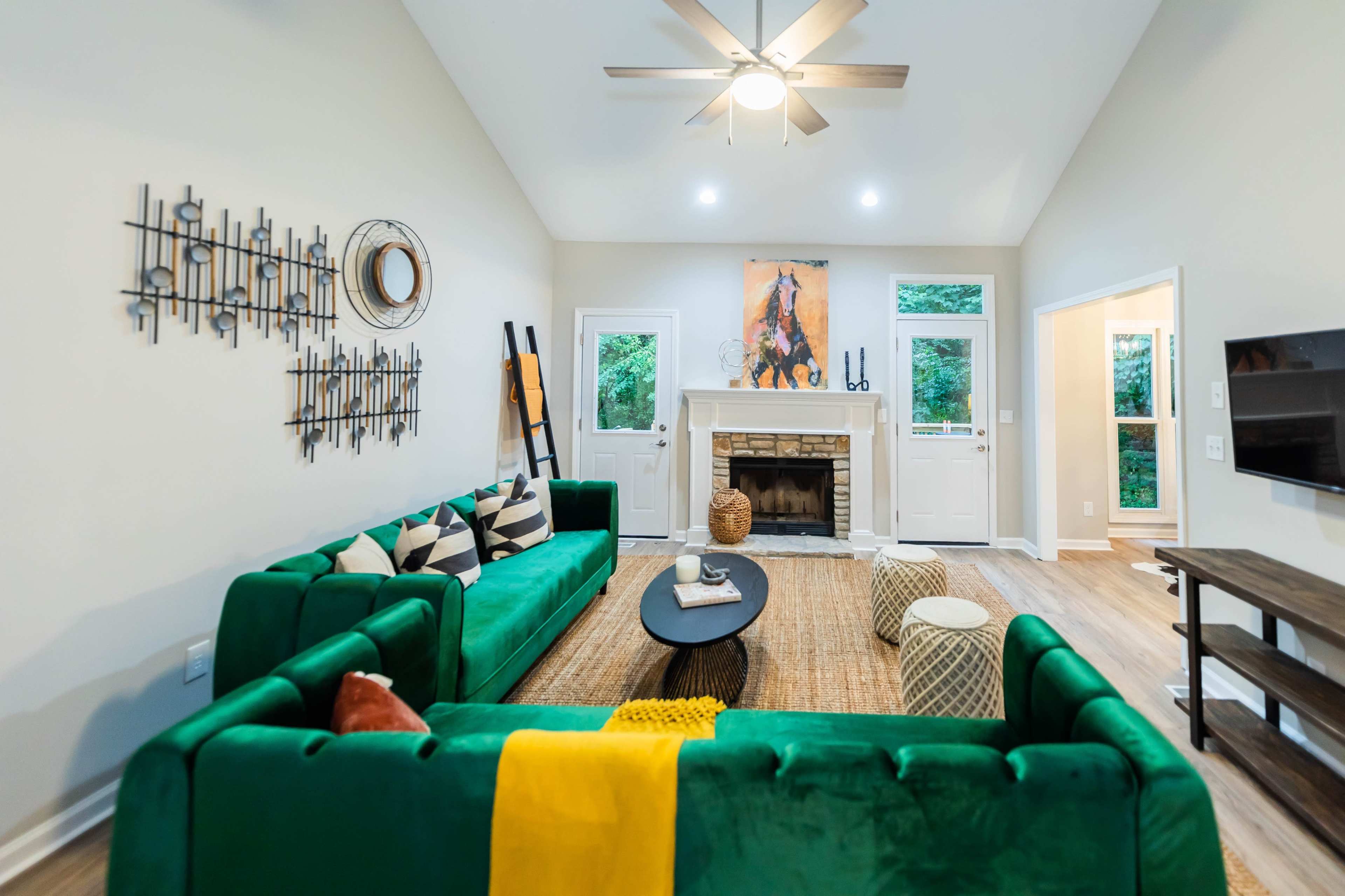 A modern living room features two green sofas facing each other, a central coffee table, a fireplace, and large windows that open to a view of greenery outside.