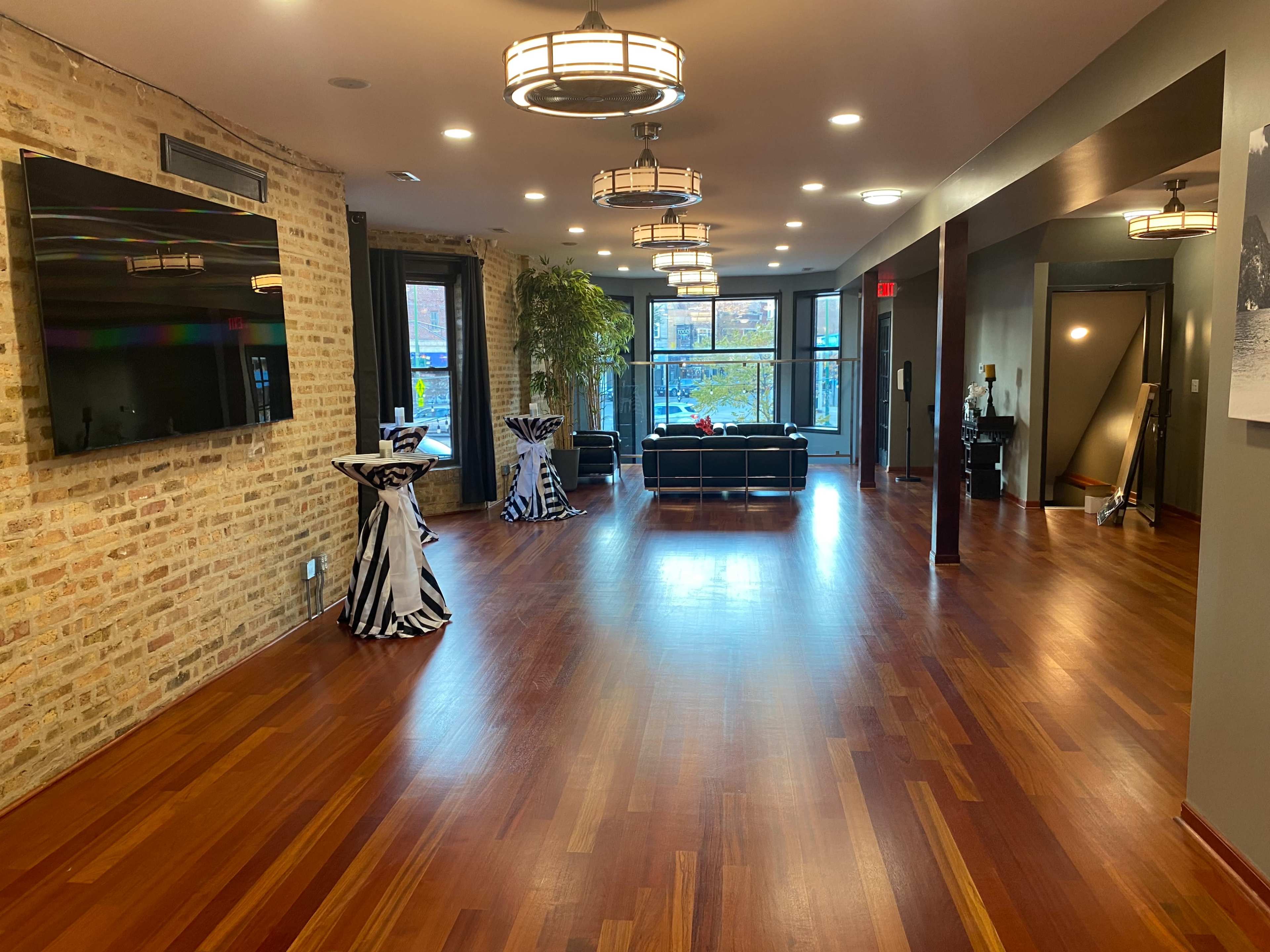 A modern interior space with wooden floors, a large TV on a brick wall, and seating arrangements, including black sofas and tall tables.