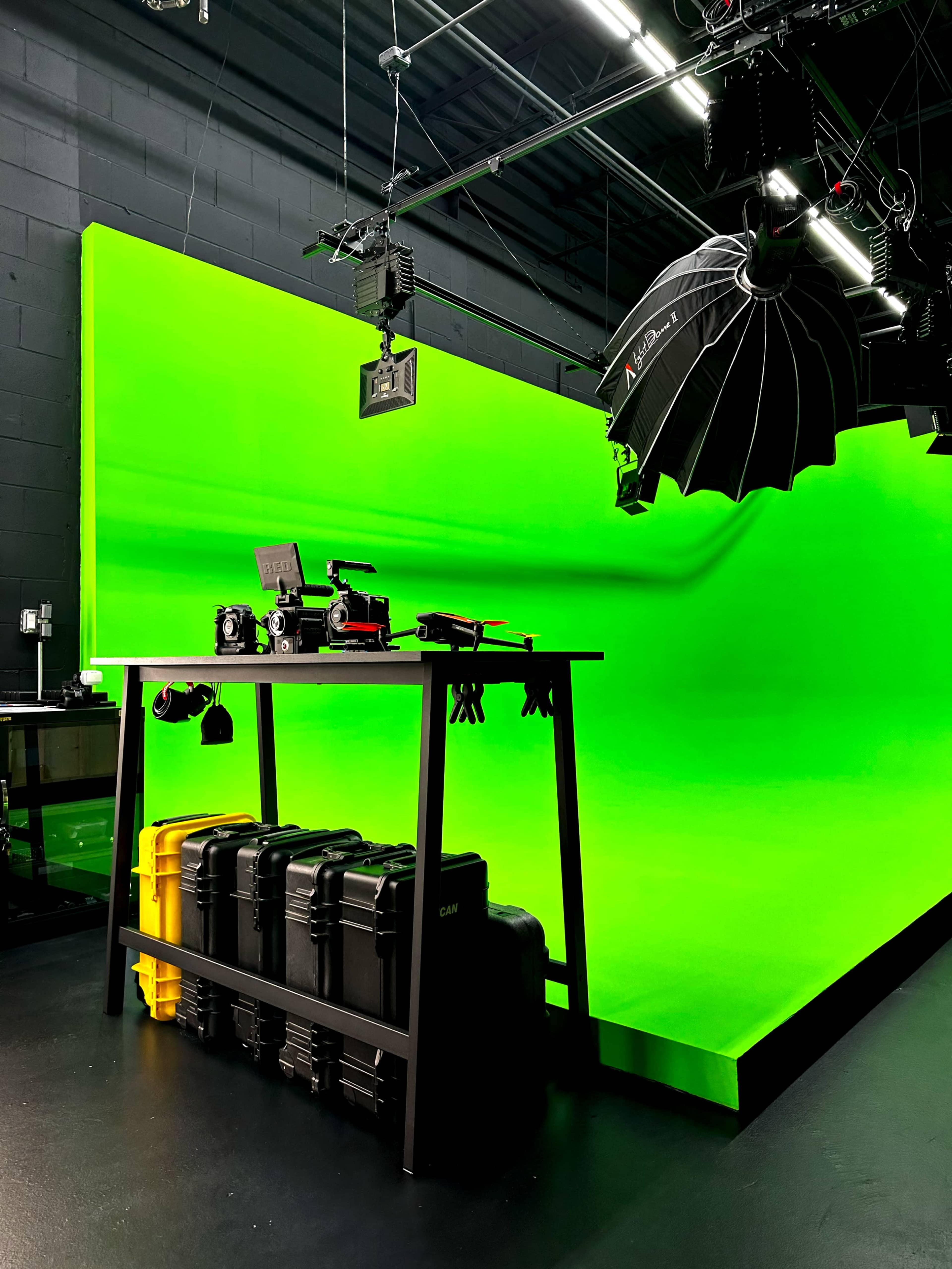 A photography studio features a green backdrop, a table with various camera equipment, and light modifiers suspended from the ceiling.