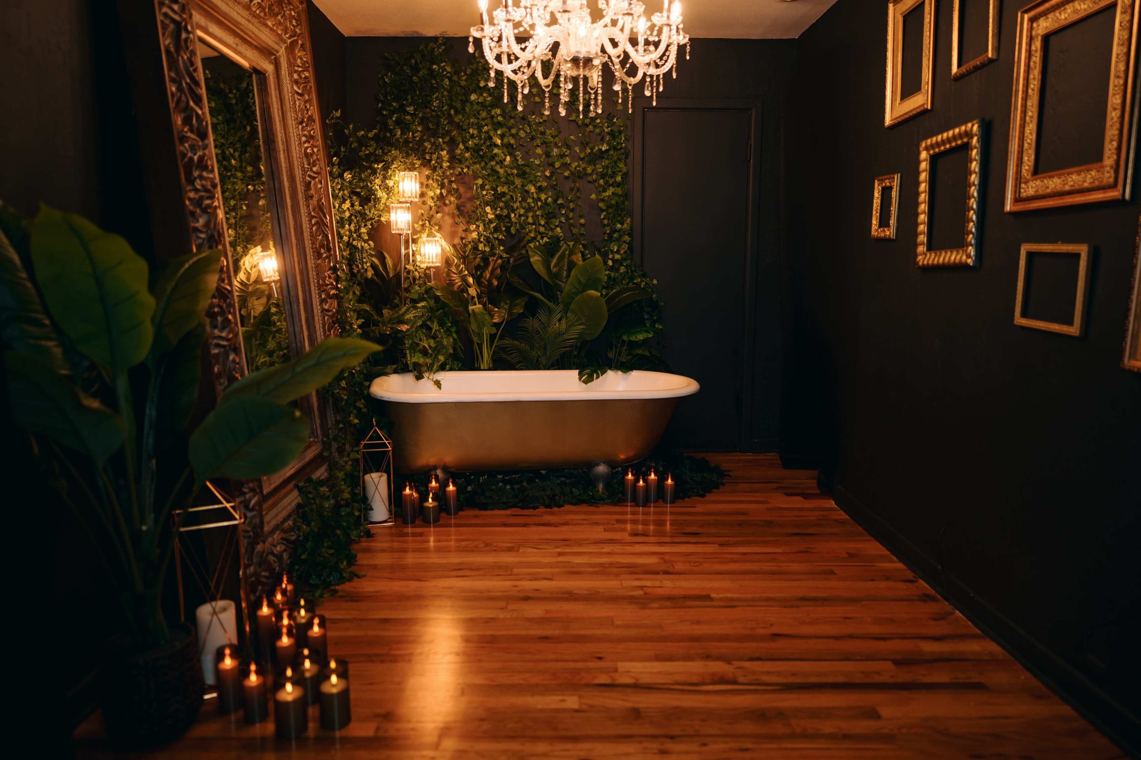 The image shows a stylish bathroom featuring a freestanding bathtub surrounded by greenery, candles, and a large ornate mirror, all under a chandelier.