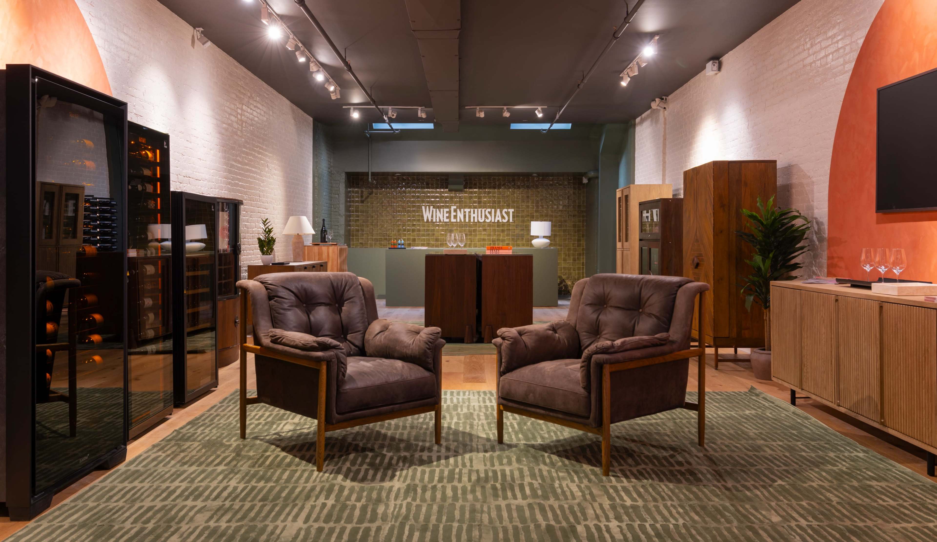 The image features a modern retail space with two upholstered armchairs facing each other on a patterned rug, surrounded by wine coolers and wooden furniture displays.