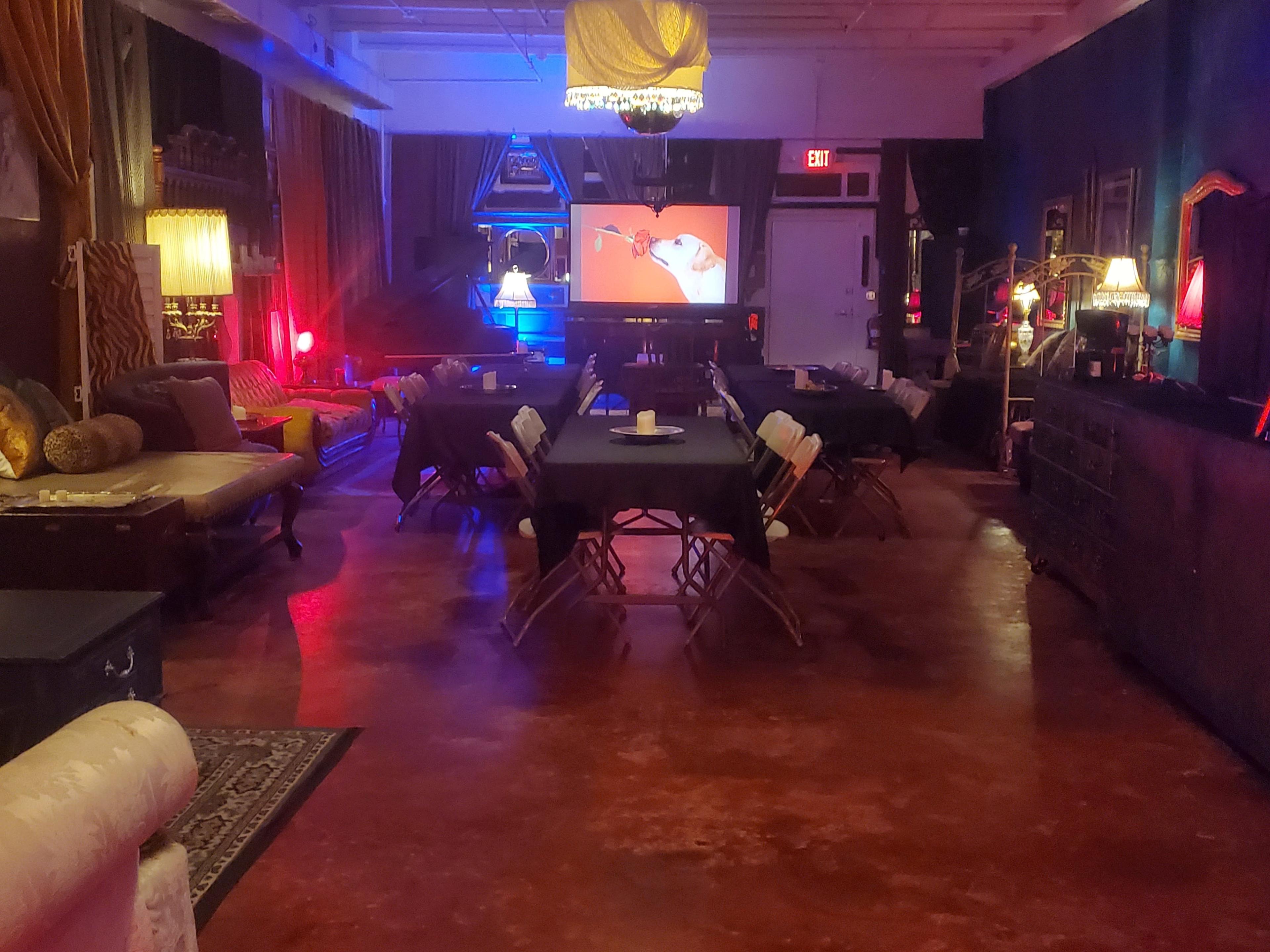 The scene shows a dimly lit event space with folding tables, chairs, and a large screen displaying a colorful image.