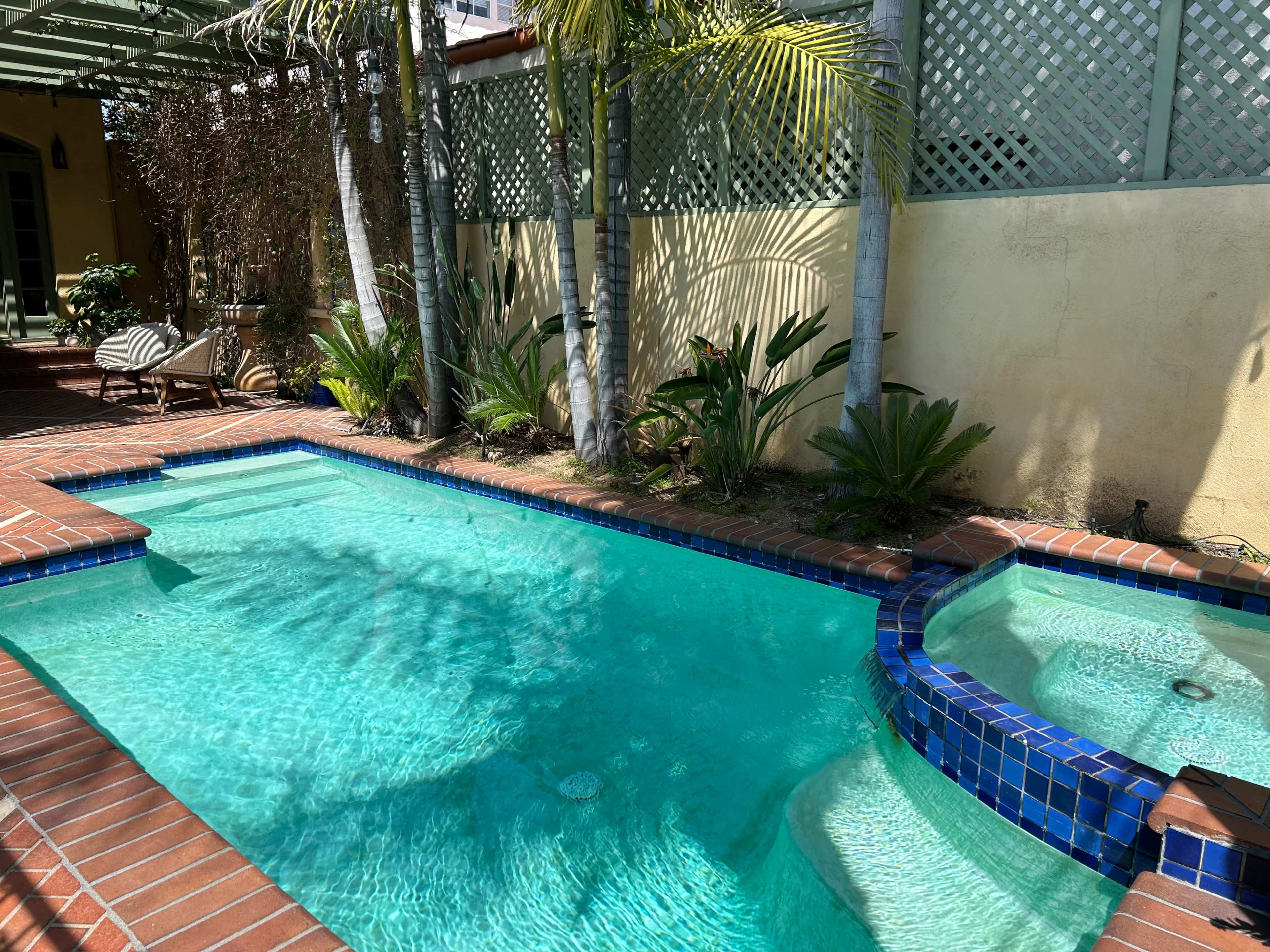 A swimming pool surrounded by palm trees and brick paving, with a built-in hot tub on one side.