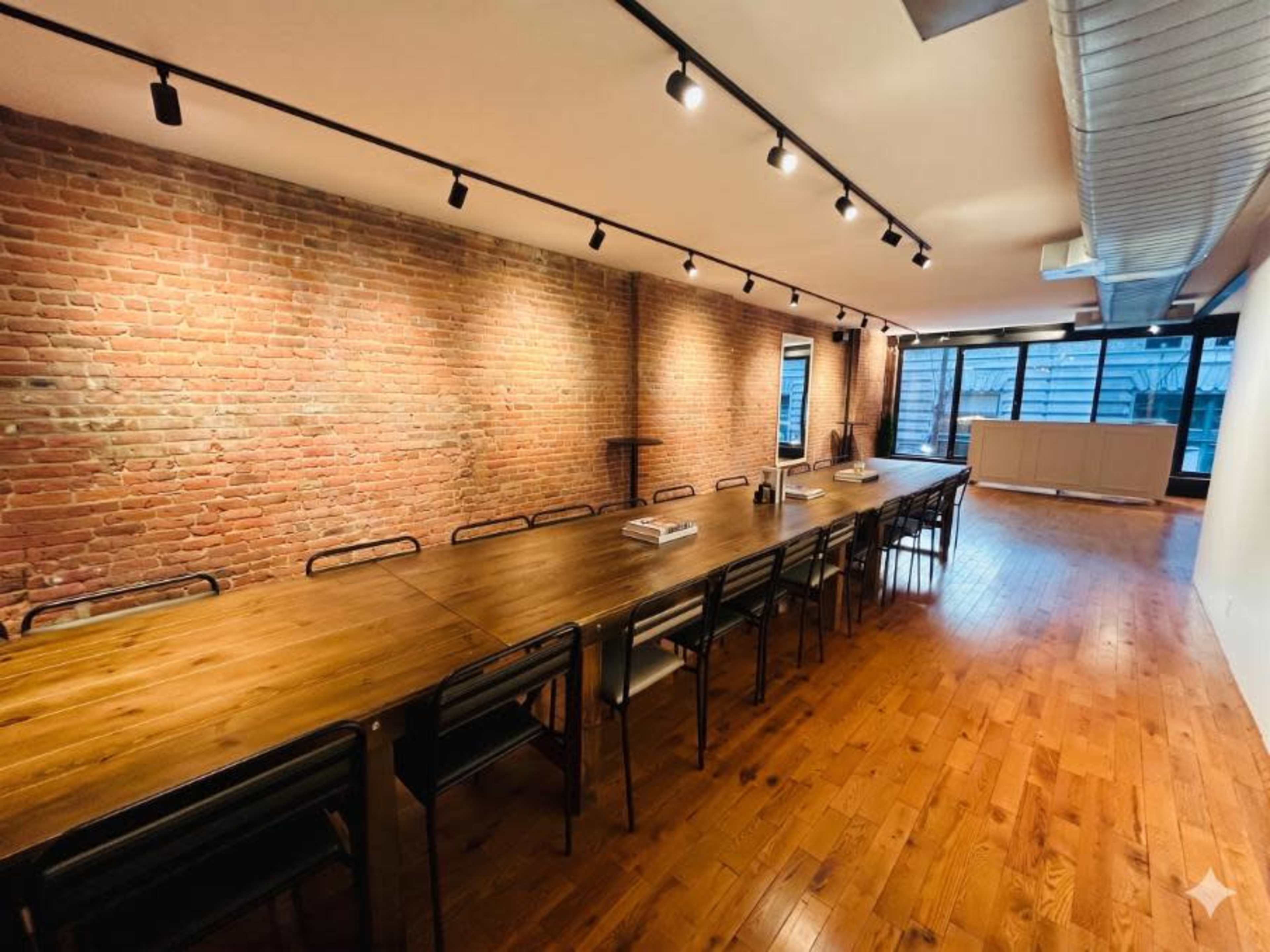 The image shows a spacious meeting room featuring a long wooden table surrounded by black chairs, with exposed brick walls and large windows allowing natural light.