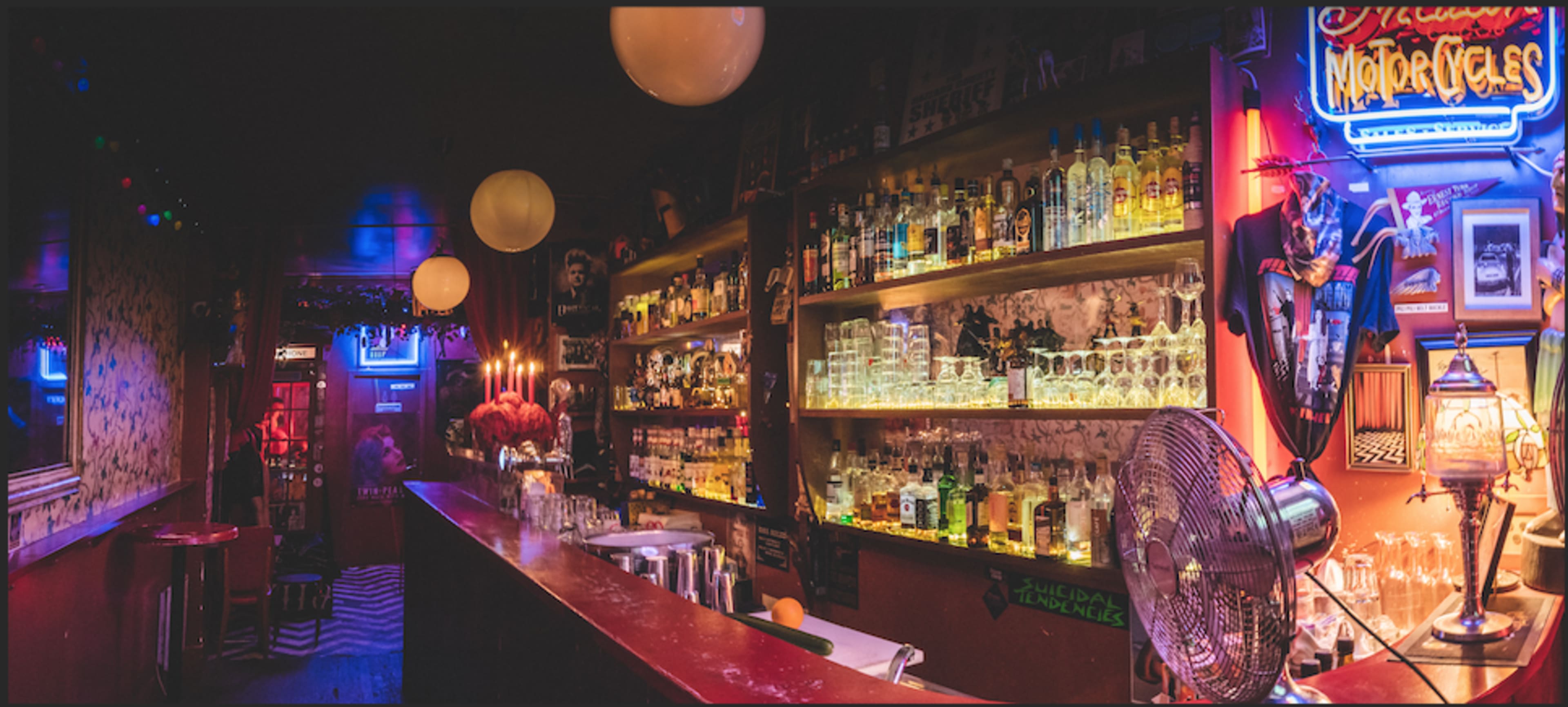 The image shows a dimly lit bar with shelves lined with various bottles of alcohol, illuminated by neon signs and decorative lighting.