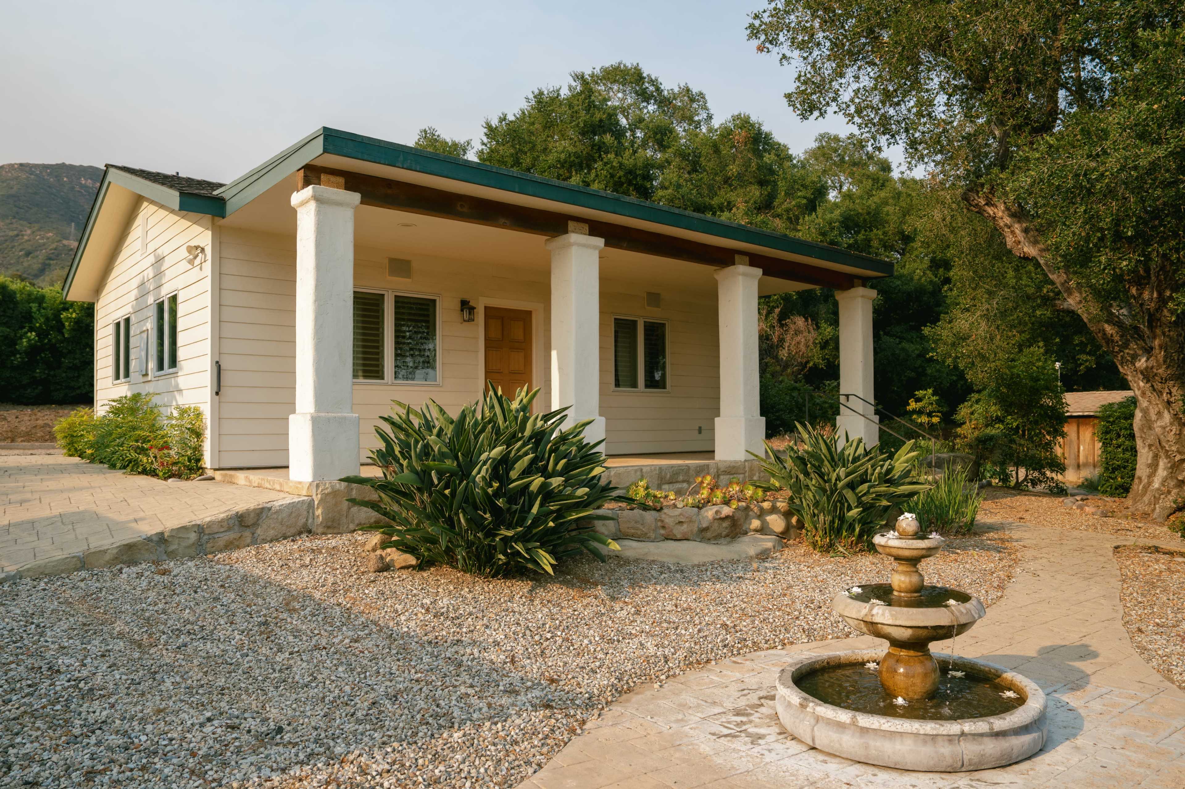 A single-story house with a front porch, surrounded by gravel landscaping and a small fountain in the foreground.