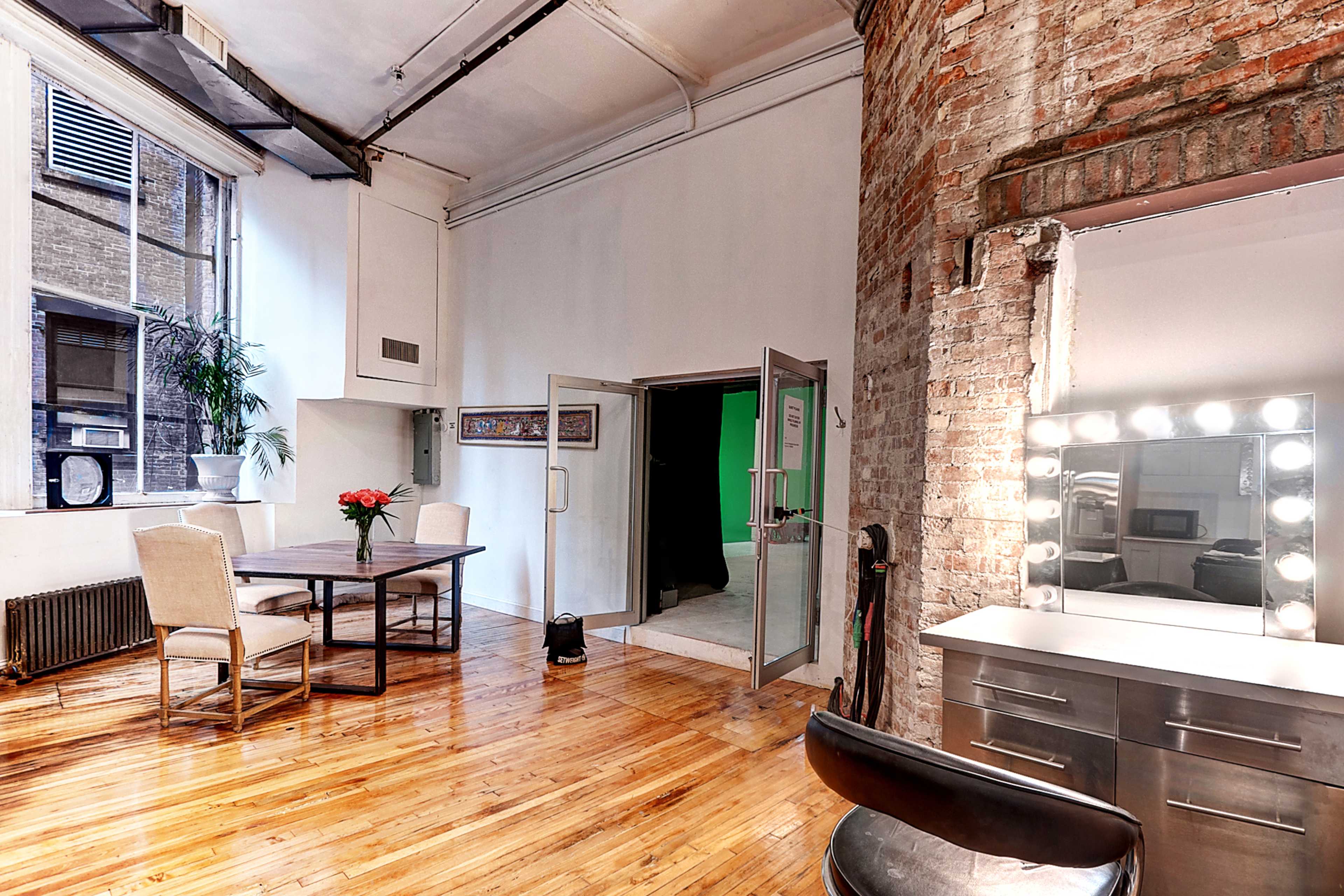 The image shows a spacious room with wooden flooring, a dining table and chairs, a vanity with lights, and an entrance to a green-screen area.