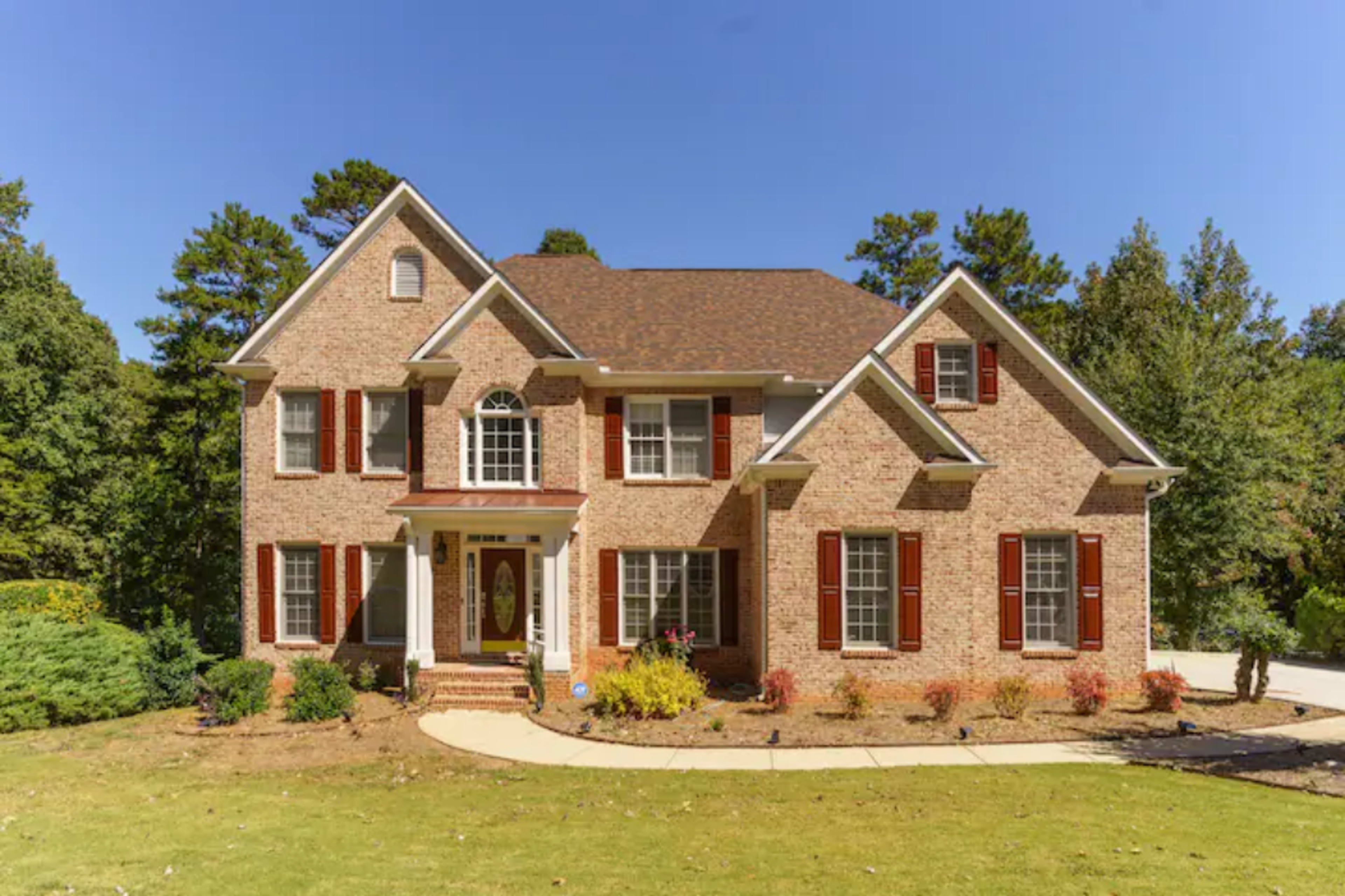 The image shows a two-story brick house with a symmetrical facade, featuring white trim and red shutters, surrounded by landscaped greenery.