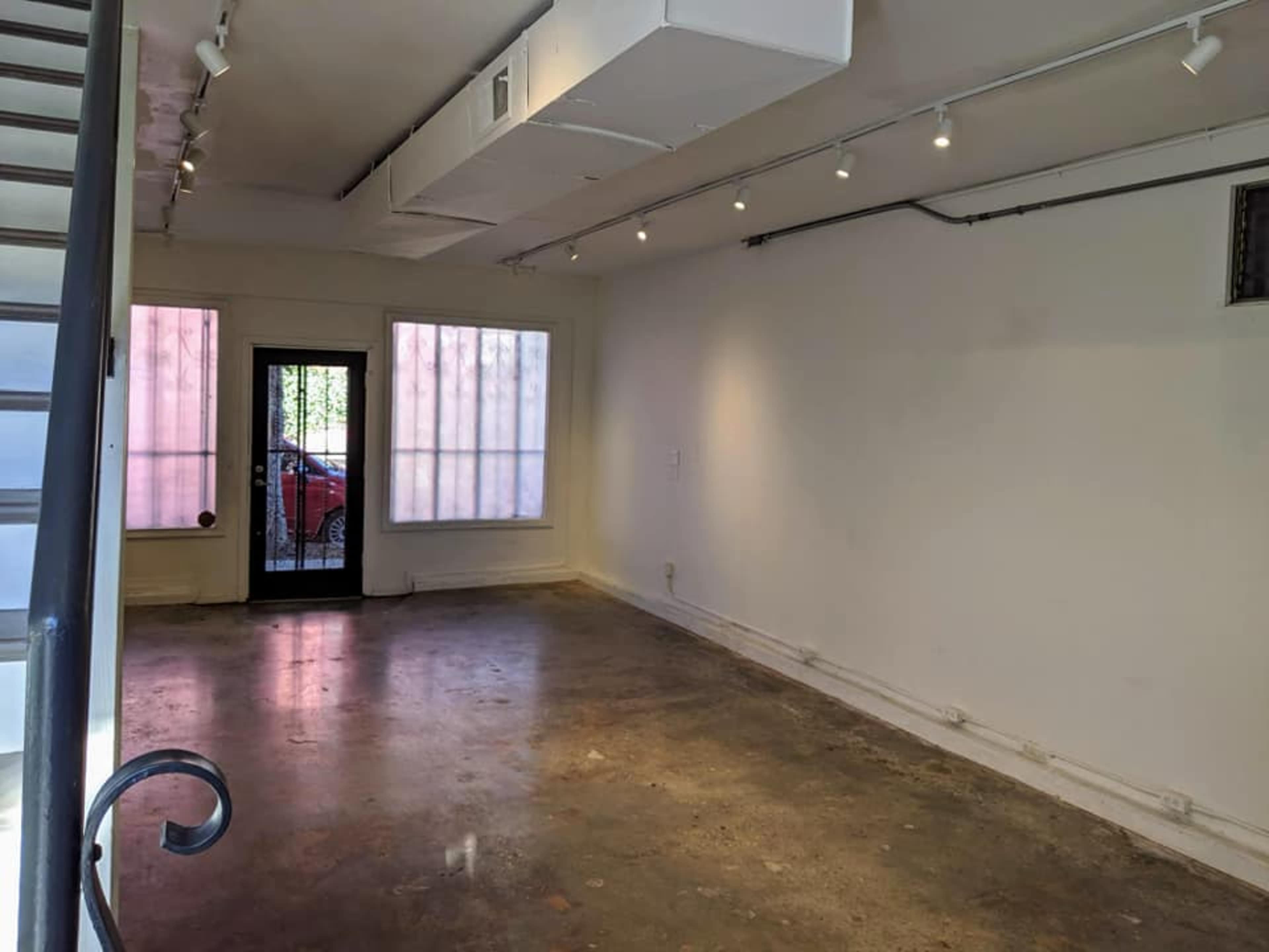 The image shows an empty gallery space with a concrete floor, white walls, and large windows on one side.