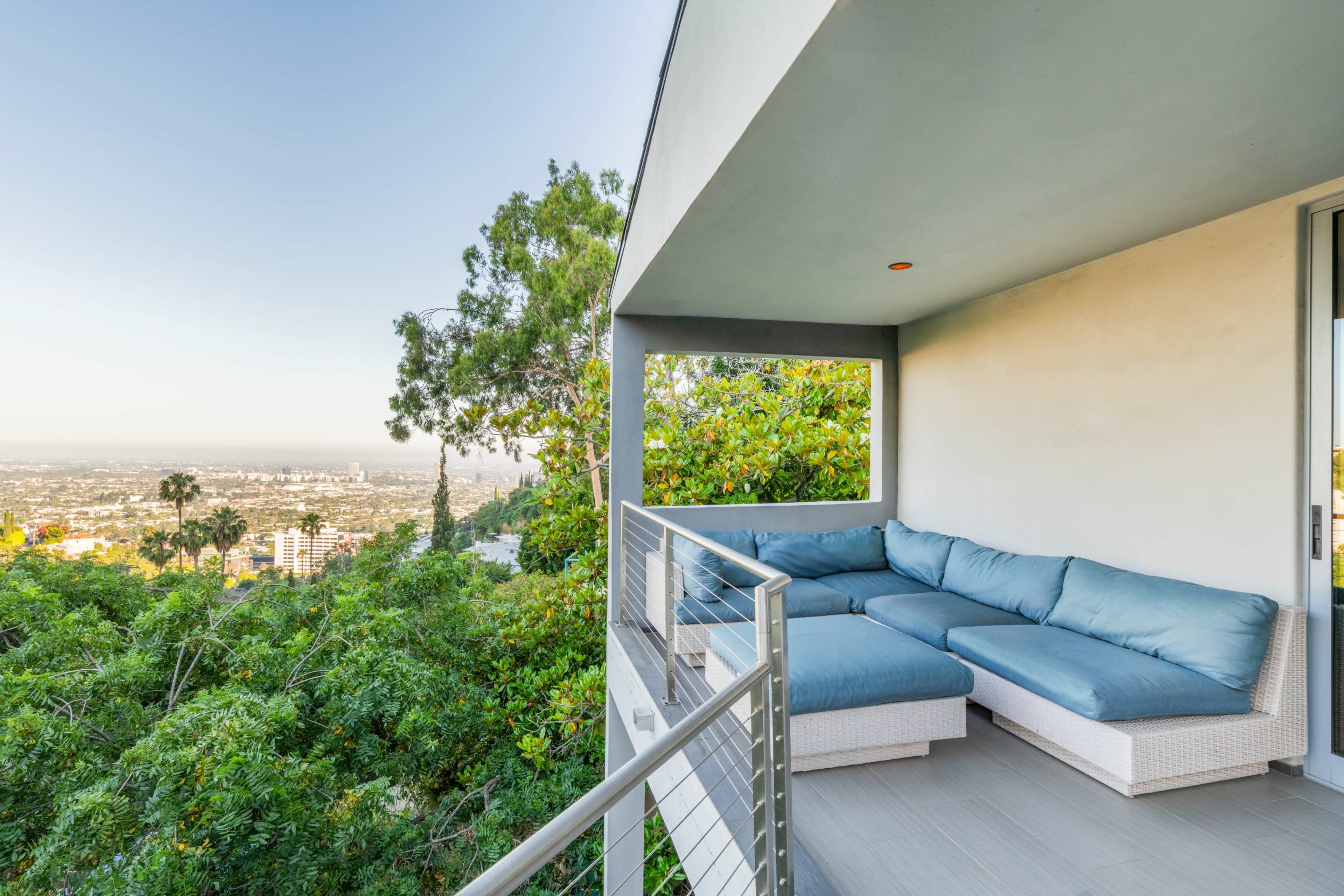 A modern balcony with a blue cushioned seating area overlooking a green landscape and city skyline.