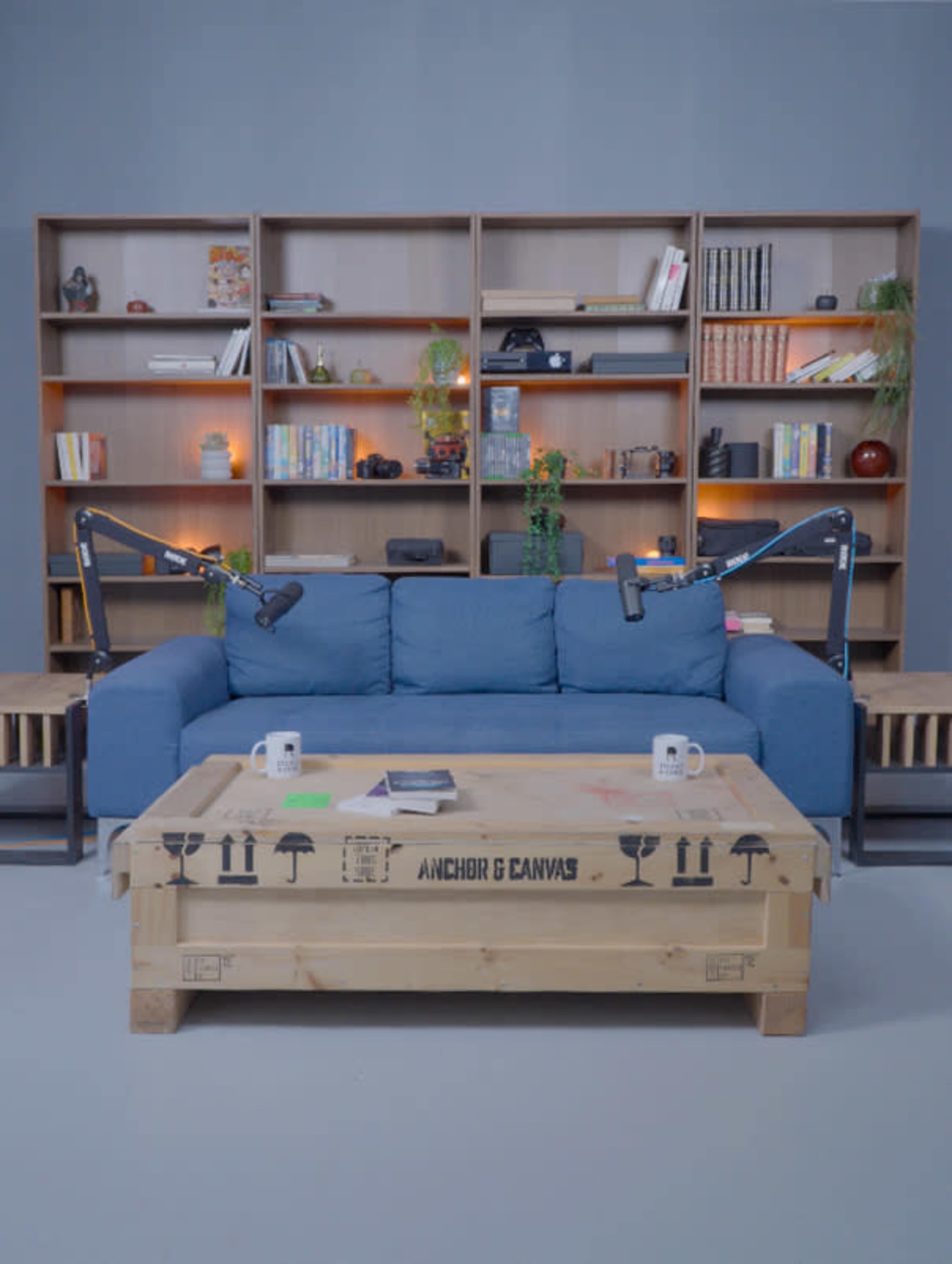 The image shows a blue couch in front of a wooden coffee table, with two microphones mounted on arms and a bookshelf filled with books and decorative items in the background.